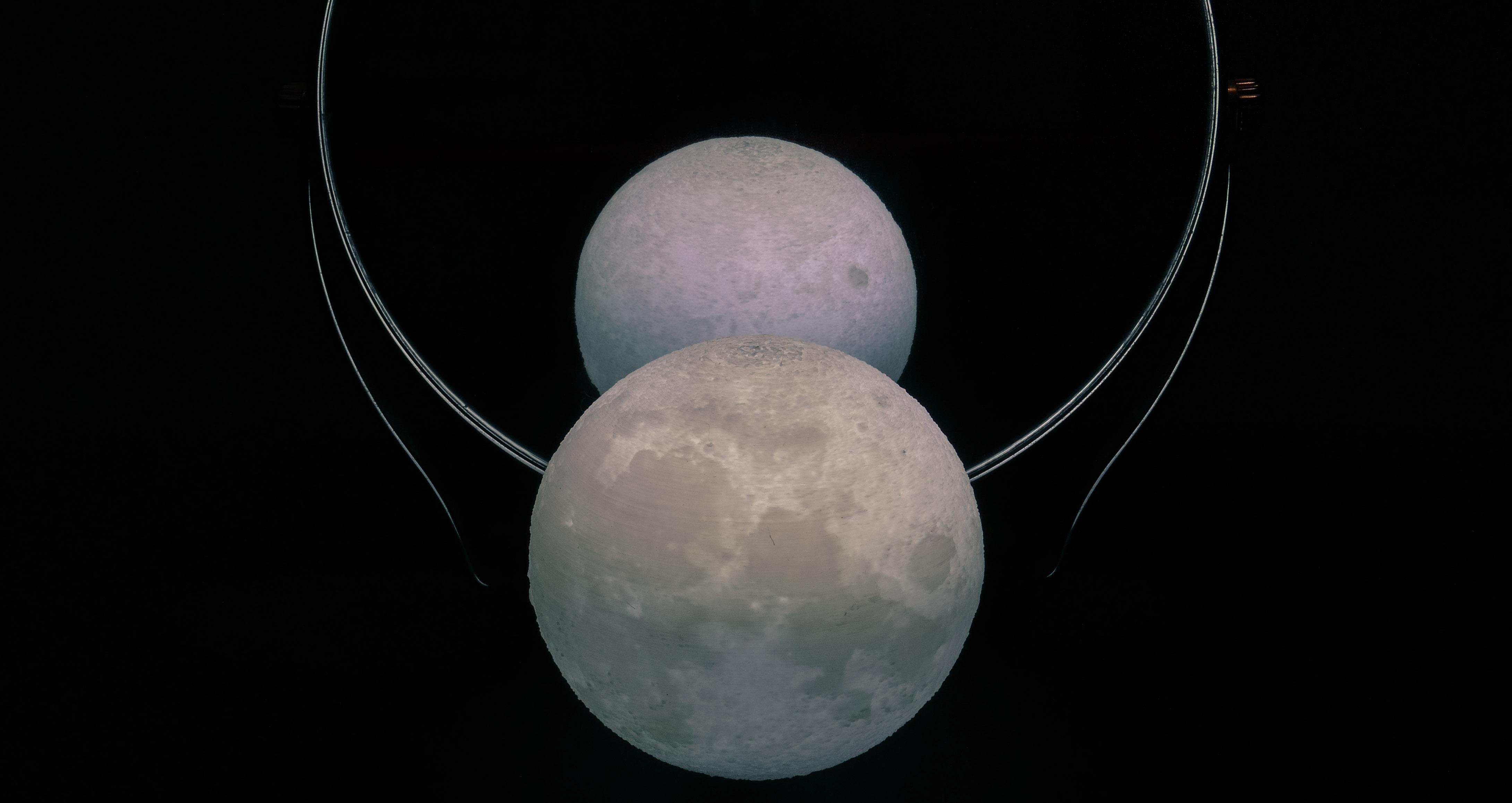 The image shows a spherical object that resembles the moon, with a reflective surface behind it creating the appearance of two moons. The reflection effect suggests the use of a mirror.