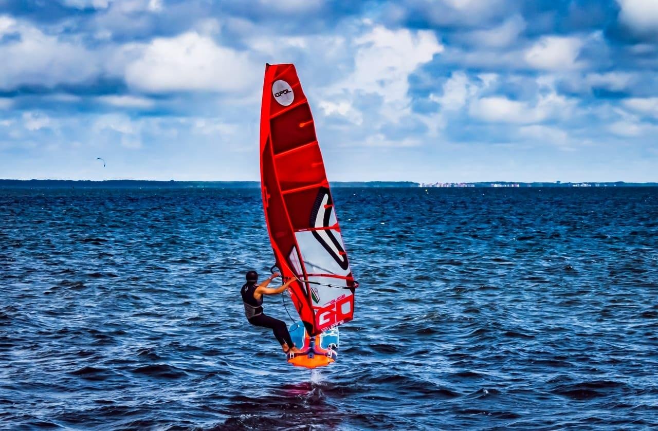 Windsurfer in a wetsuit riding a board with a large red sail on choppy blue water, with a distant shoreline on the horizon under a cloudy sky.