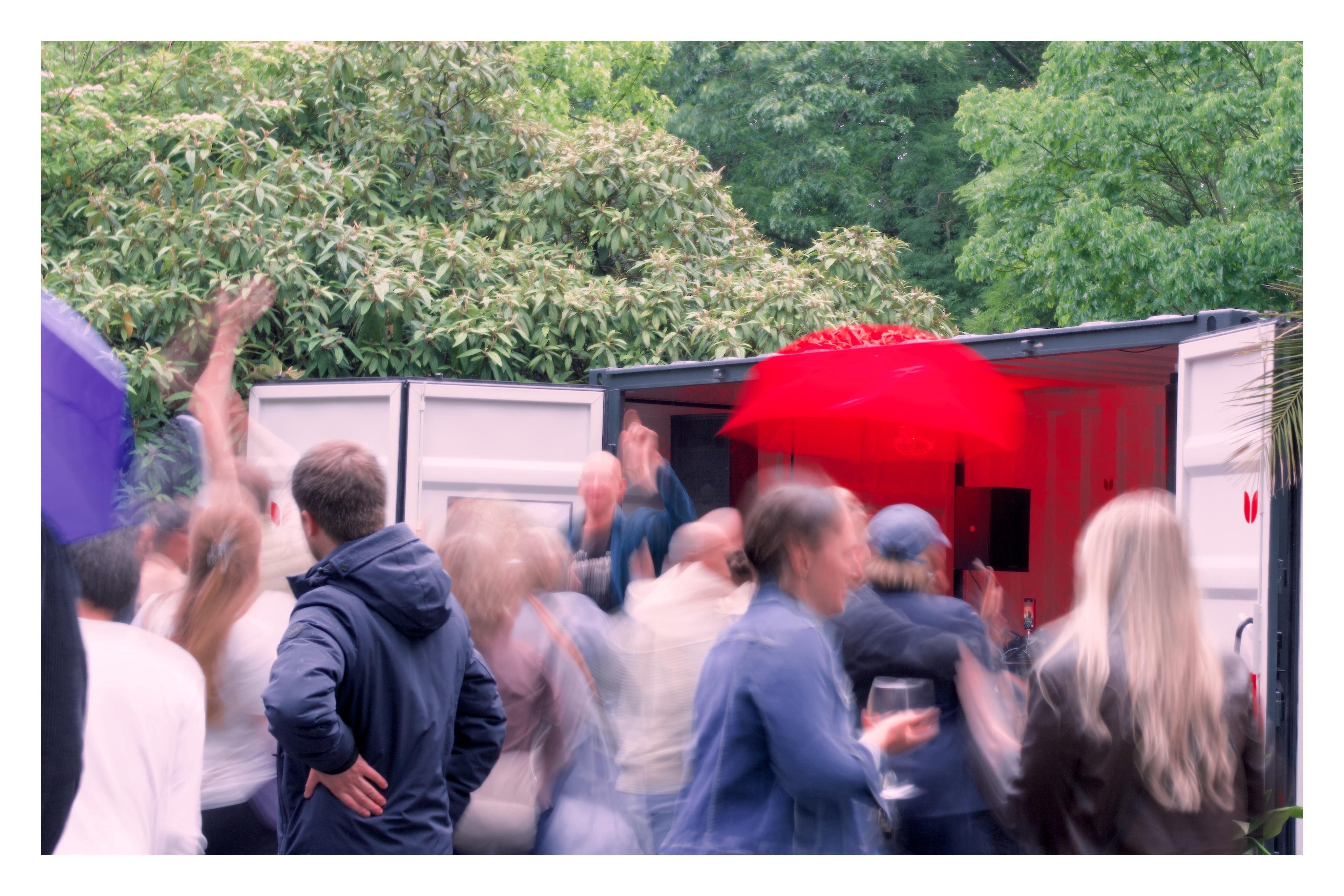 Gruppe von Menschen feiert vor einem geöffneten Container im Grünen, einige Bewegungen verschwommen, rote Beleuchtung im Container.
