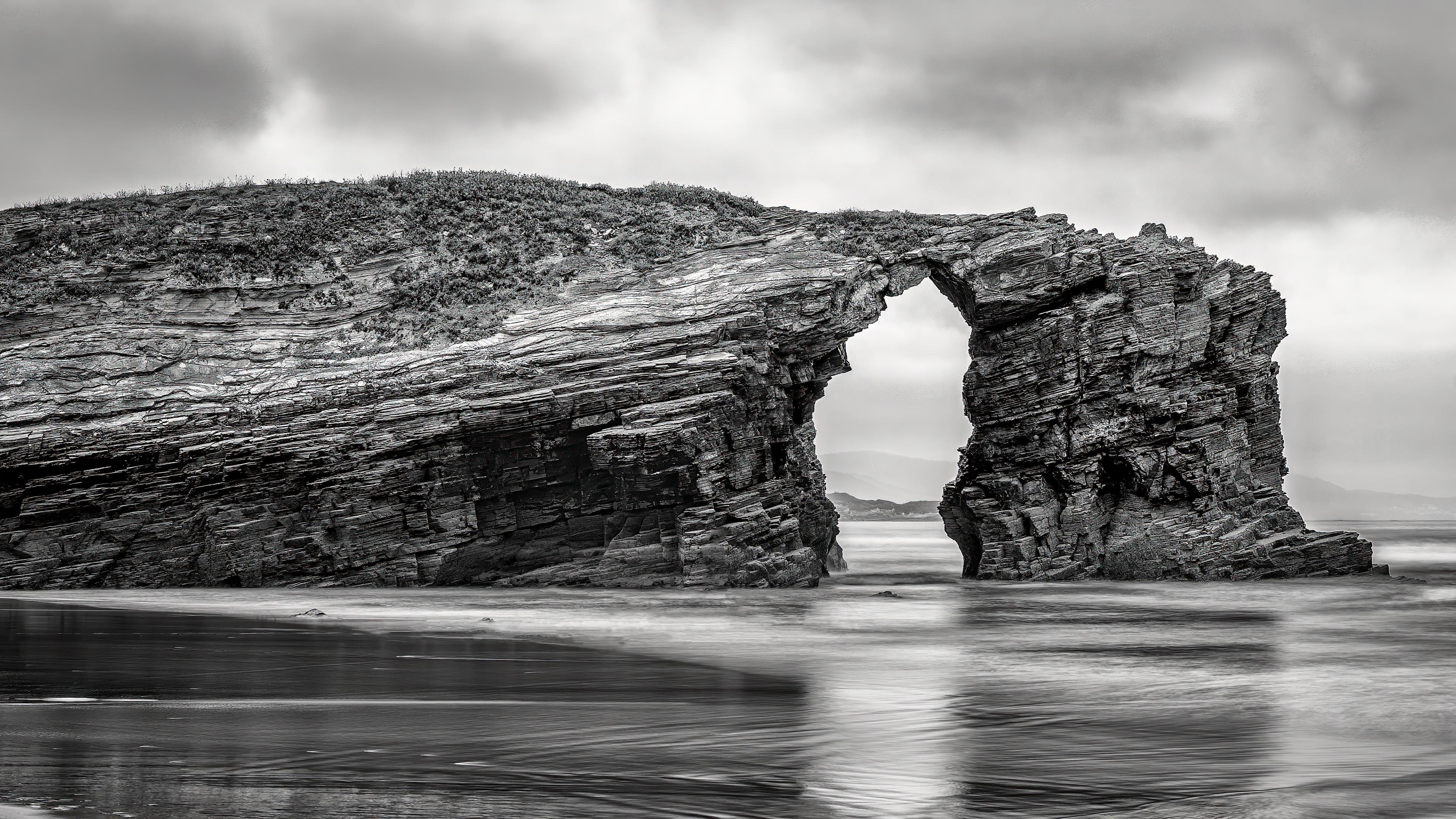 Rock formation with a natural arch over the sea, appearing in grayscale. The rocks have layered striations, and the tide is low, revealing wet sand in the foreground. Cloudy sky above adds a moody atmosphere.