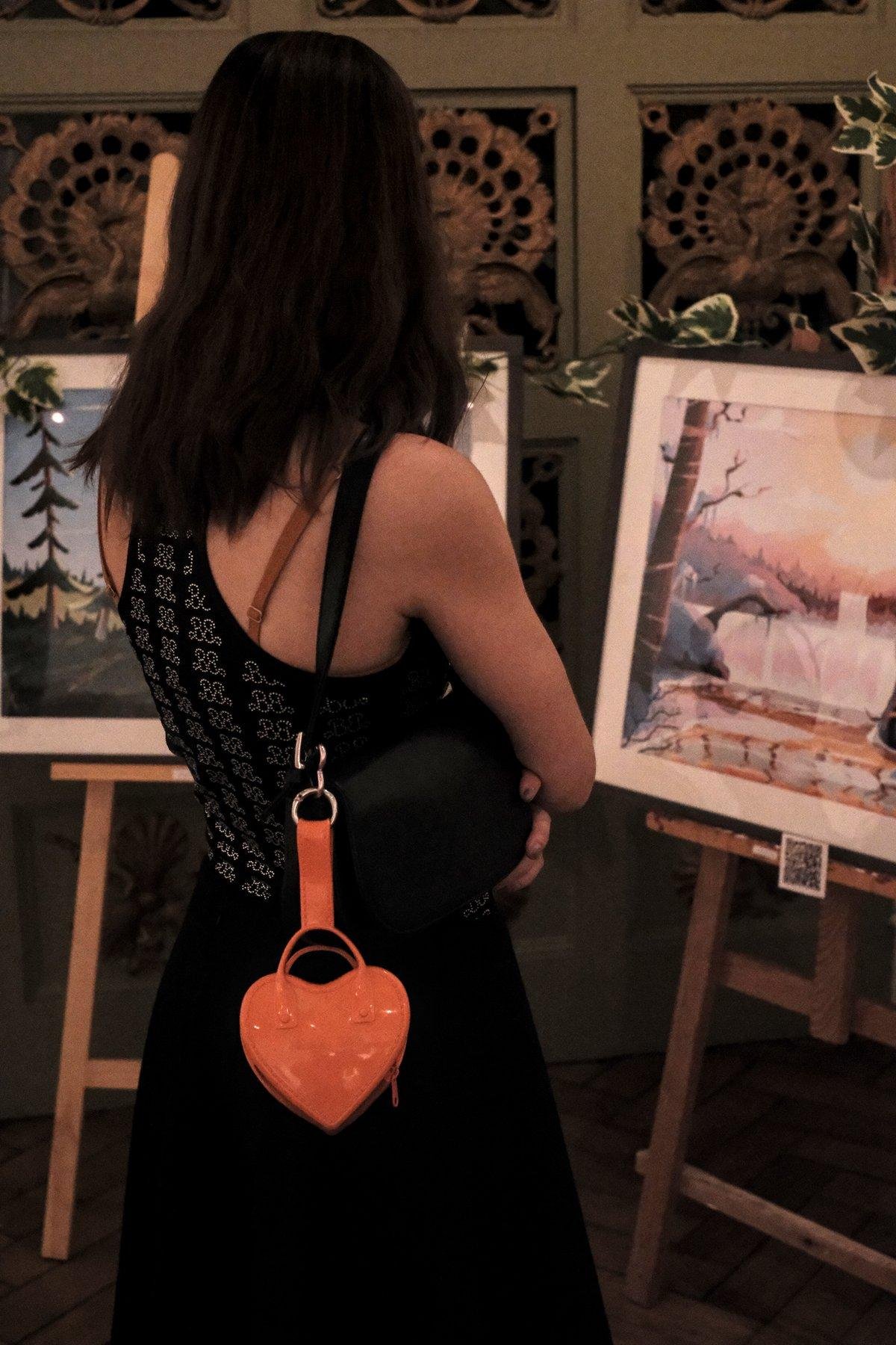 Photo verticale. Une jeune femme en haut sans manche noir à motifs faits d’une série de couples de lettre B dont celle de droite est à l’envers pour faire face à celui de gauche, faits en petit brillants, jupe longue noir, et petit sac à main en forme de cœur orange. Elle regarde des tirages d’œuvres numérique