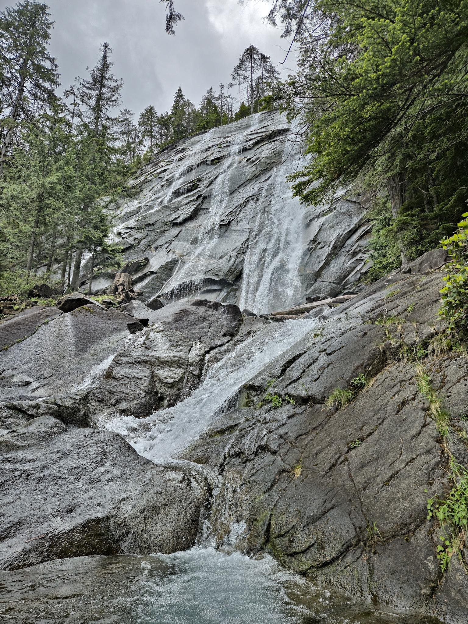 Waterfall cascading down a steep rocky cliff surrounded by lush green trees. The water flows vigorously over dark, jagged rocks before settling in a pool below. The sky above is overcast, adding a soft, diffused light to the scene.