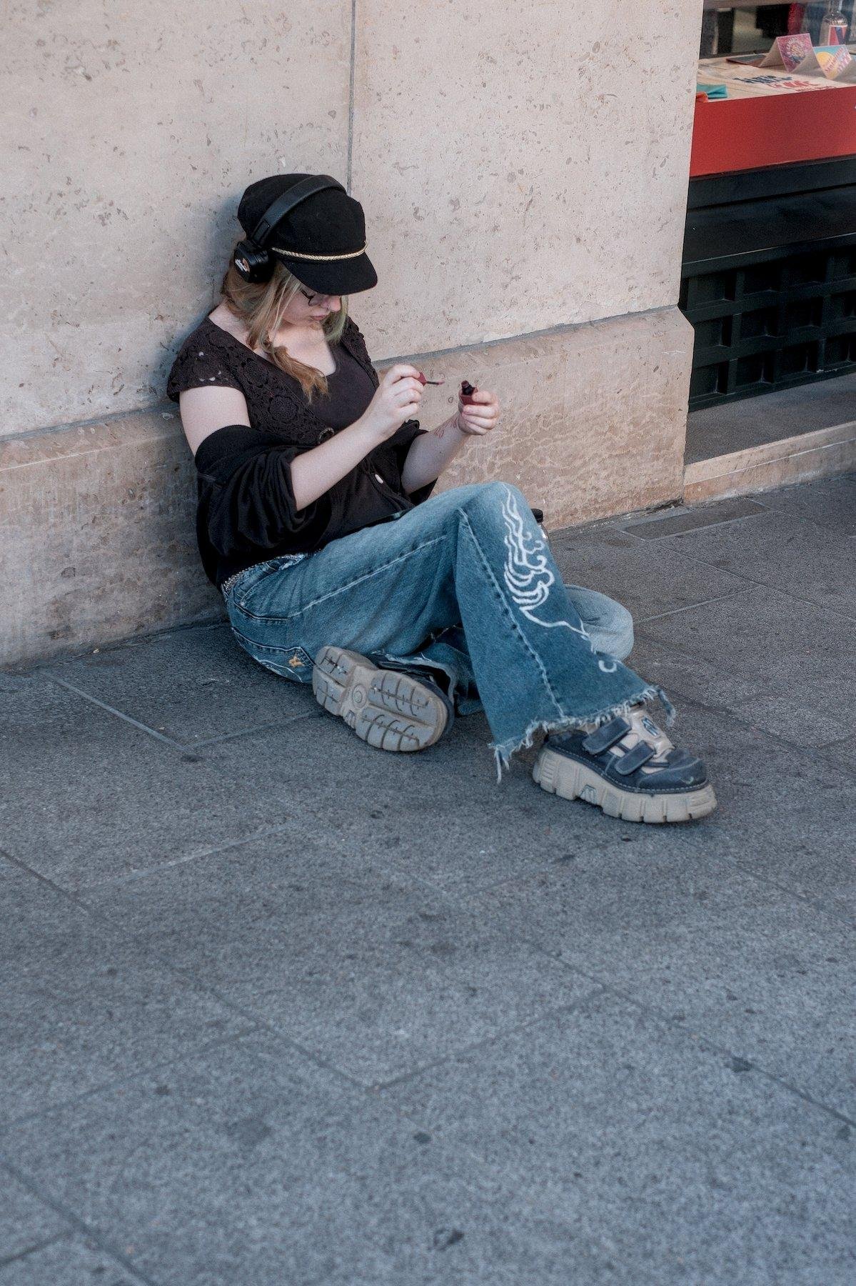 Photo verticale. Une jeune femme châtain en jean avec une broderie stylisé sur le tibia, chaussures à grosses semelles gris clair, haut noir, casquette et casque audio sur la tête, concentrée sur ses ongles qu’elle est en train de vernir, assise sur le trottoir, adossée contre un mur