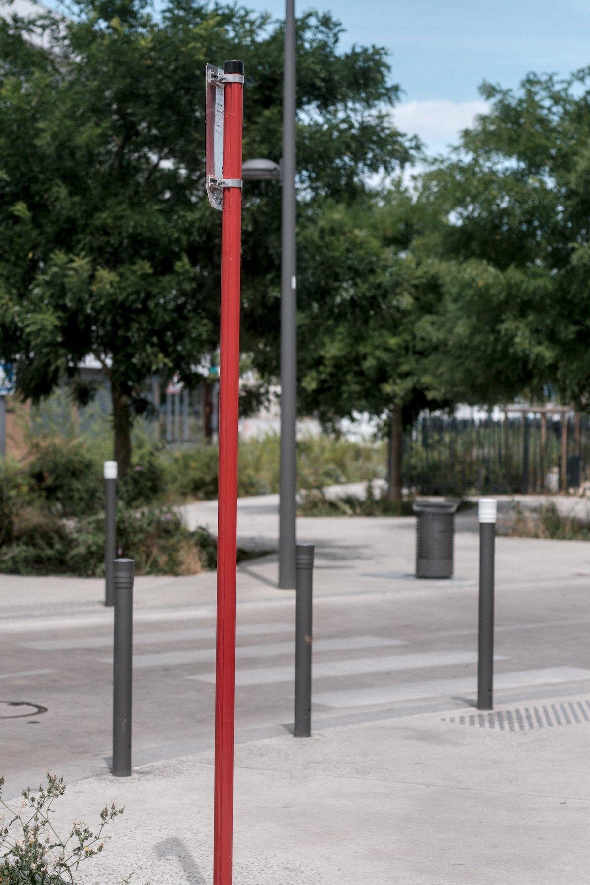 Photo verticale. Dans la rue, un poteau rouge ressort du reste de l’environnement majoritairement beige, gris et vert