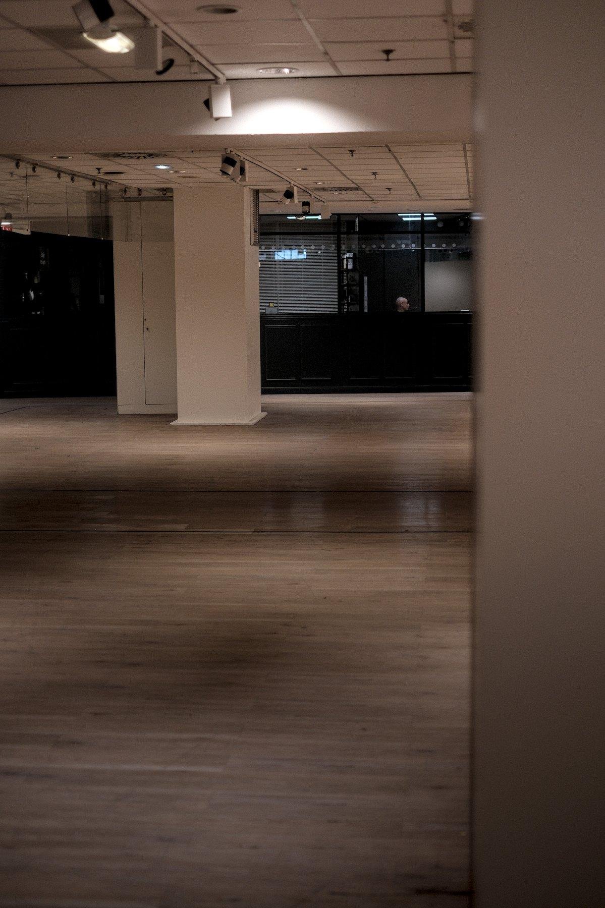 Photo verticale. Un grand espace au parquet vide. Des colonnes carrées tiennent l’espace, et au fonfd, une délimitation gris sombre, avec de grandes baies vitrées, et soudain, la tête de profile d’un homme au crâne rasé dépasse d’une vitre, en train de parler à quelqu’un qu’on ne voit pas.