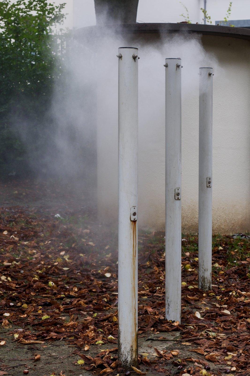 Photo format portrait de 3 poteaux blancs usés avec des coulures de rouilles qui sont en fait des brumisateurs en train de fonctionner, une brume massive s’étant formée autour d’eux. Des feuilles mortes au sol et un bout de petit bâtiment au mur blanc sale derriere eux.
