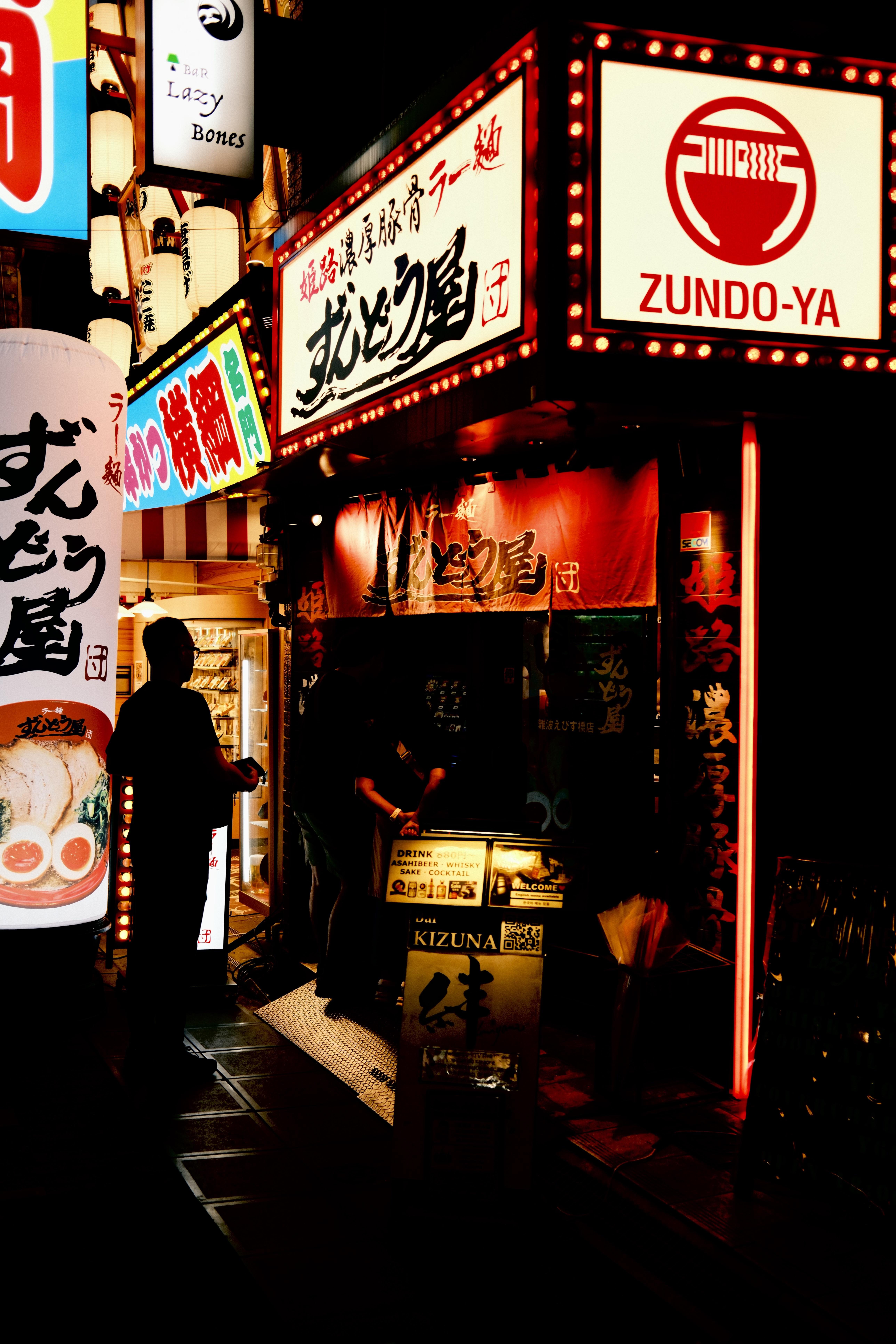 Nighttime street scene with multiple illuminated Japanese restaurant signs; large red-and-white “ZUNDO-YA” sign with ramen bowl icon above a narrow entrance draped with a red curtain, while silhouetted pedestrians pass in the foreground.