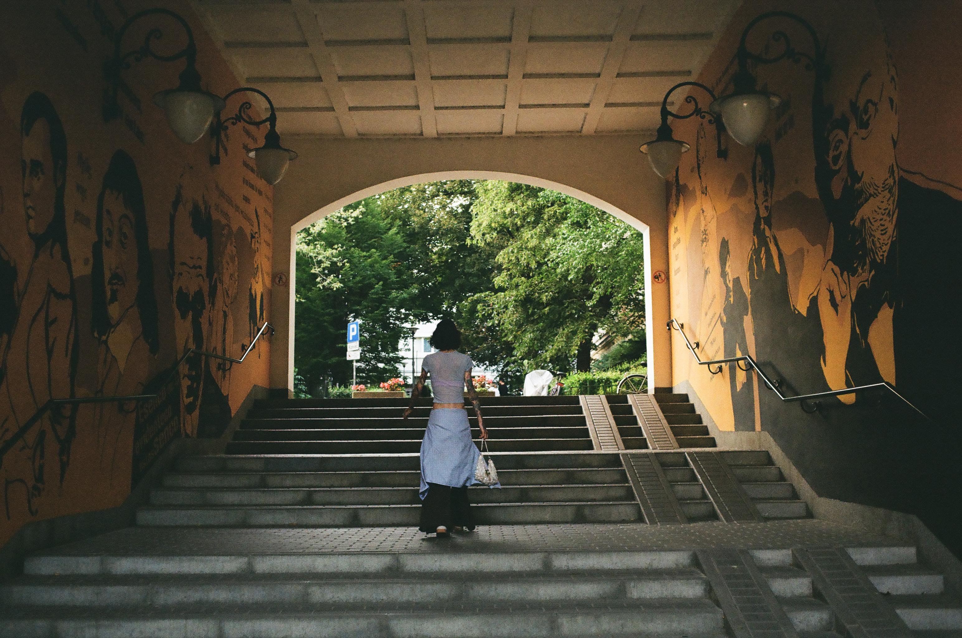 Person walking up a wide stairway through an arched underpass, with mural portraits on both side walls, metal handrails and wall lamps, and a ramp beside the steps leading toward a tree-lined street outside.