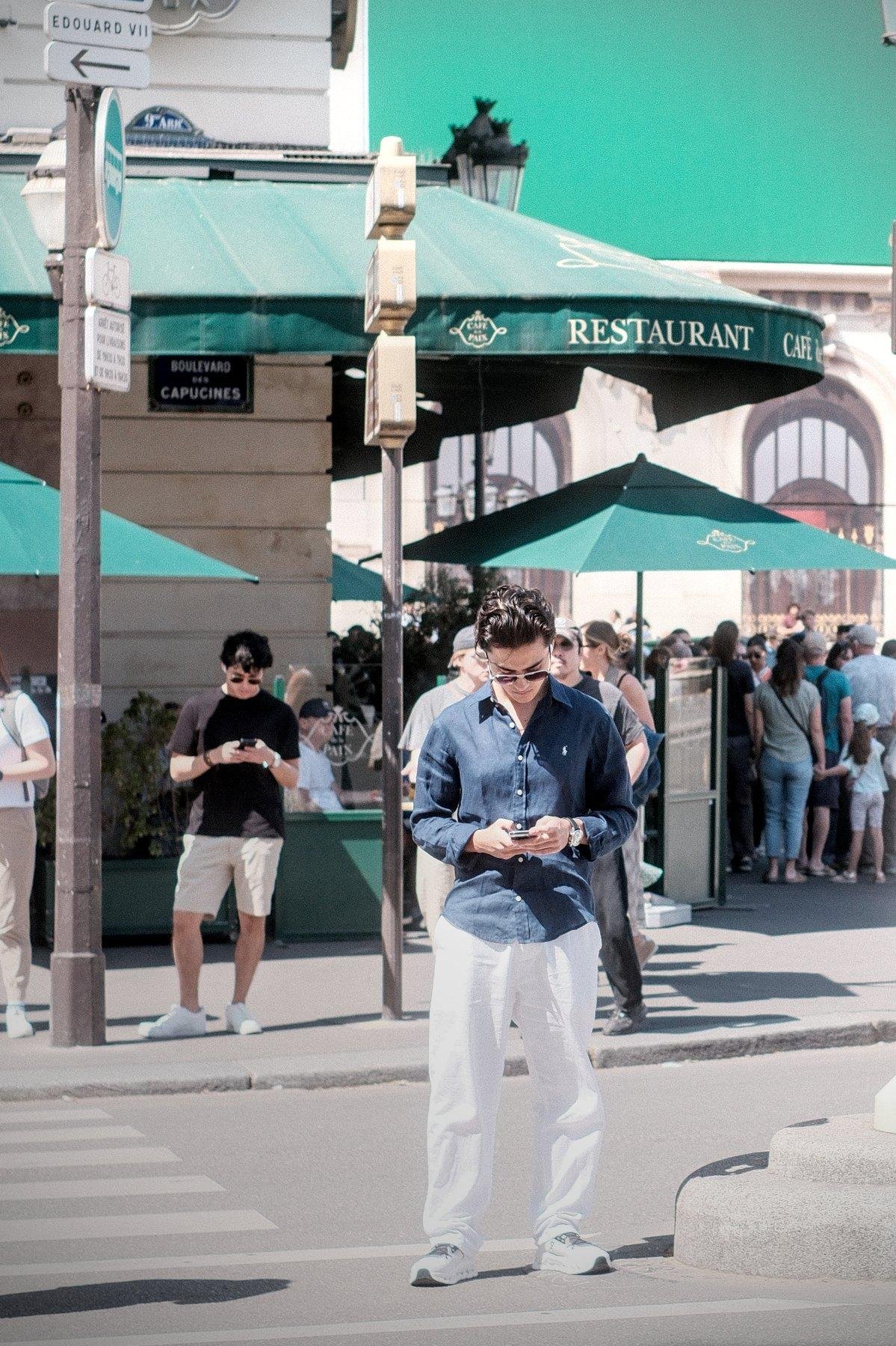 Photo verticale. Sur l’îlot de protection au centre d’une 4 voies, un homme en pantalon blanc, basket blanches, chemise couleur denim pas mise dans le pantalon, lunettes de soleil, cheveux noirs gominés, consultant son téléphone en attendant de traverser