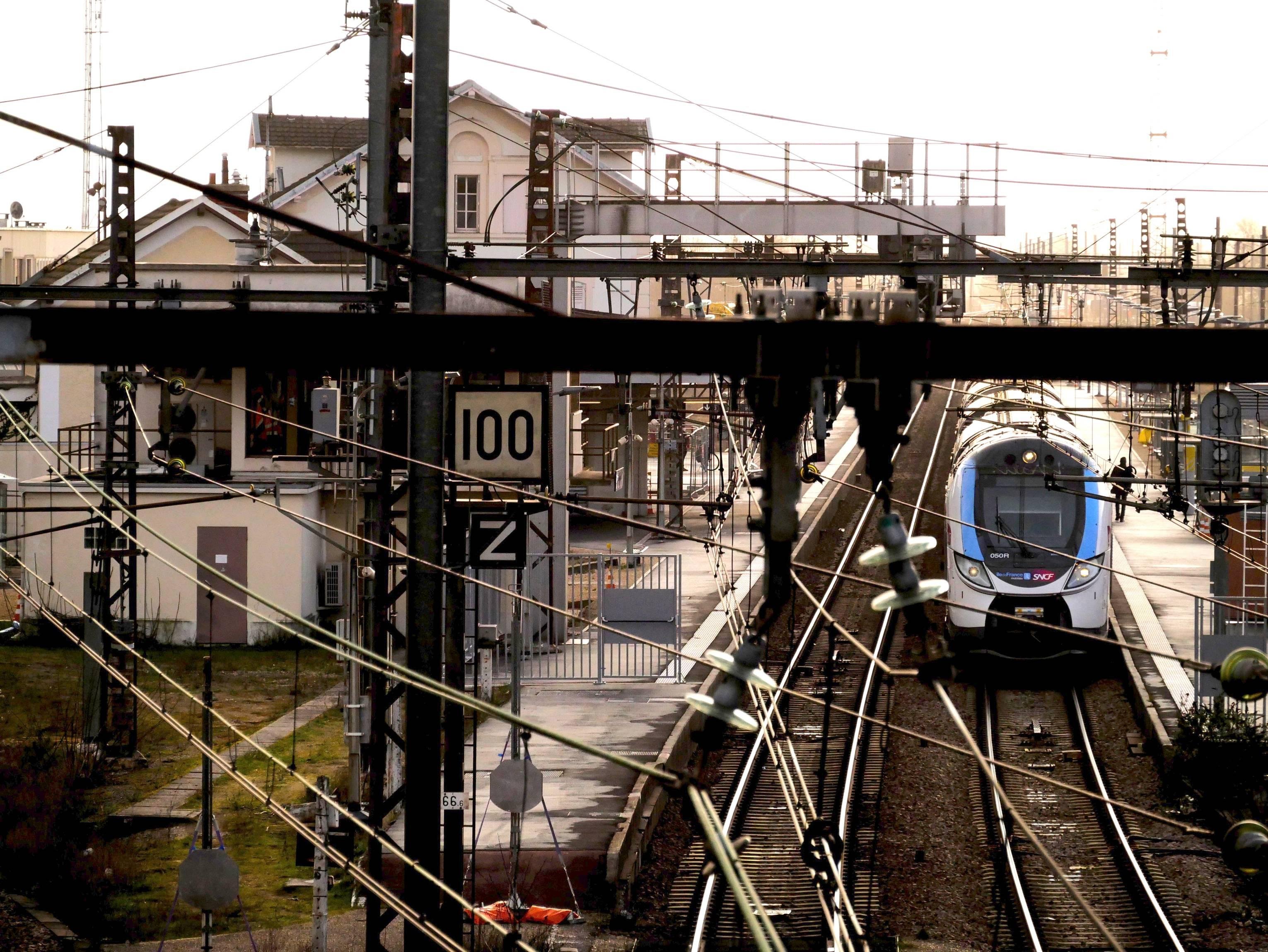The image shows a train station with a modern passenger train on the tracks. There are overhead power lines and infrastructure surrounding the area. The setting appears to be suburban. The signage visible includes a speed limit sign displaying "100" and what appears to be a directional or platform indicator marked with a "Z".