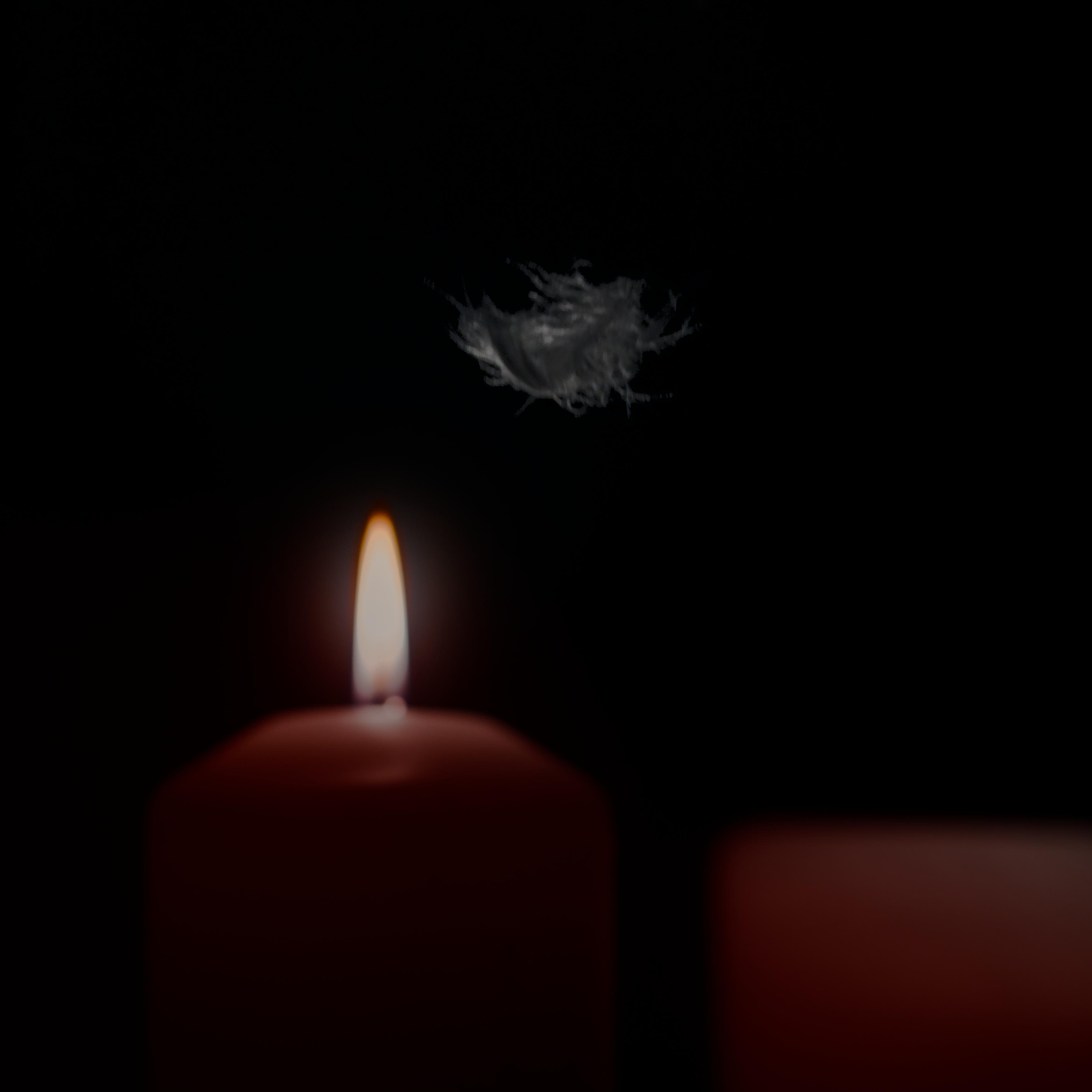 Lit candle with an orange flame on the left against a dark background, accompanied by a small, white feather floating above it. Another out-of-focus object is partially visible in the lower right corner.
###
Links ist eine brennende Kerze mit orangefarbener Flamme vor dunklem Hintergrund zu sehen, darüber schwebt eine kleine, weiße Feder. In der unteren rechten Ecke ist ein weiteres, unscharfes Objekt teilweise erkennbar.