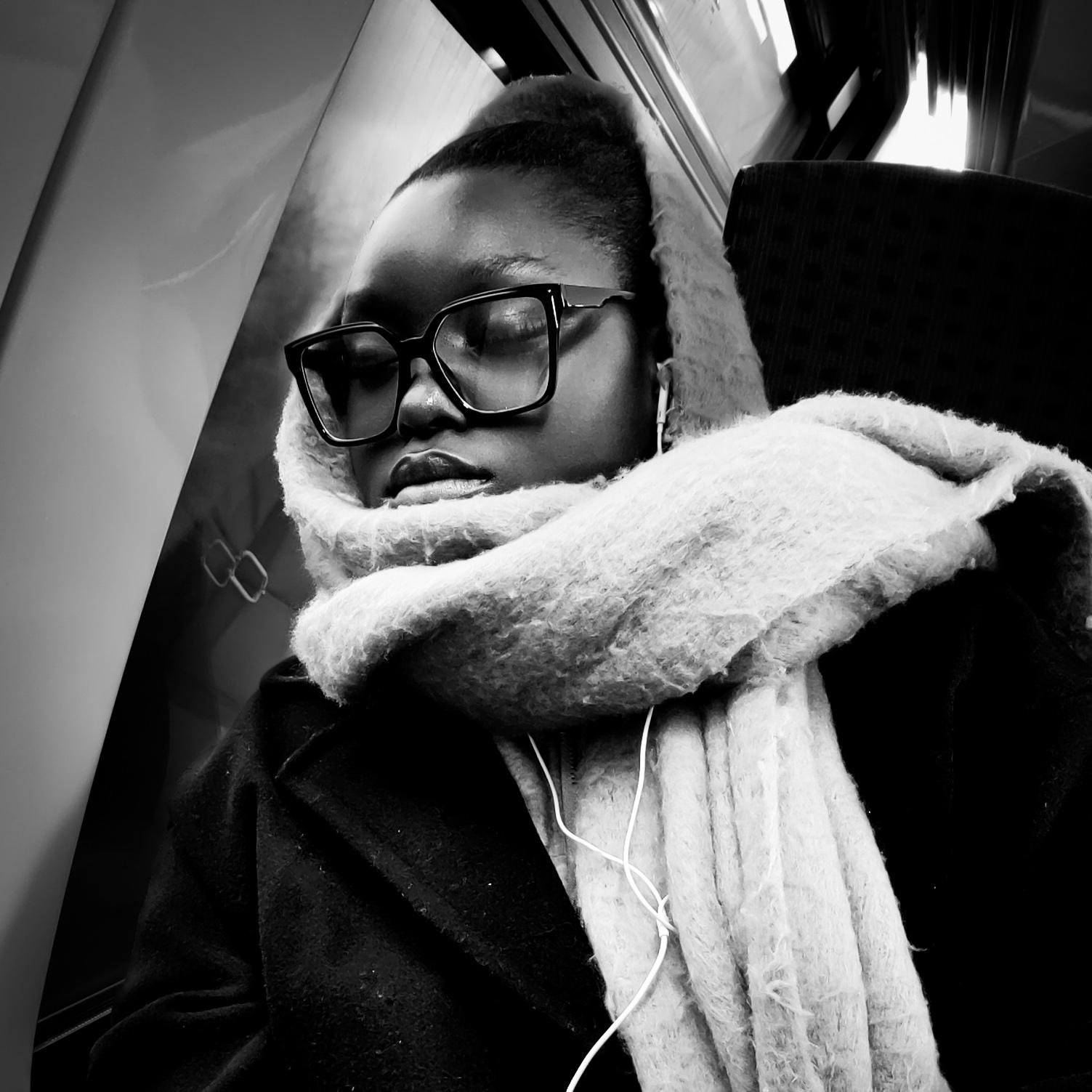 Il s'agit d'une image en noir et blanc d'une personne portant des lunettes, une grande écharpe et des écouteurs. Elle a la tête appuyée contre la vitre dans un train, les yeux fermés.