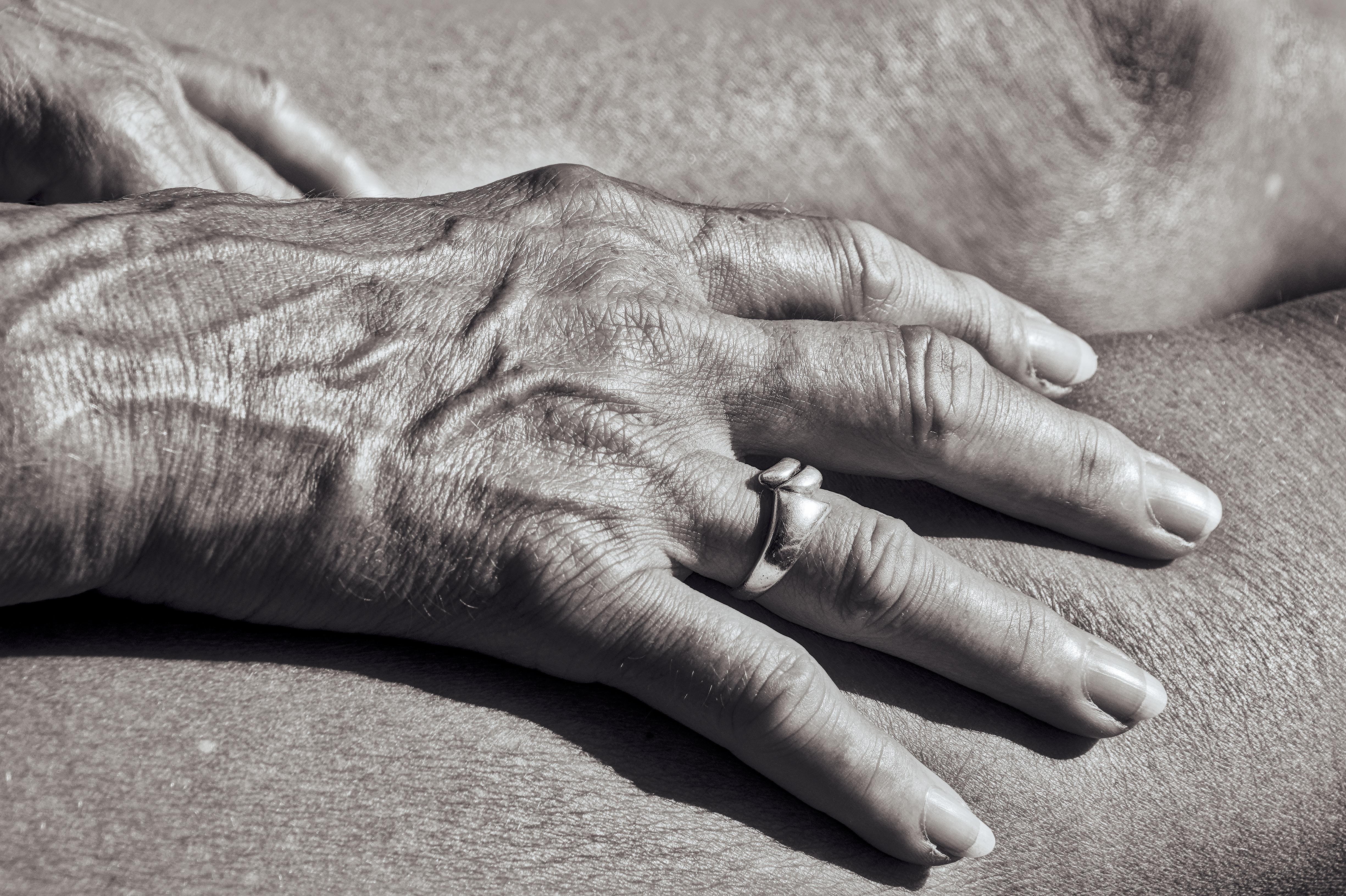 Close-up of an older person’s wrinkled hand wearing a ring, resting on another person’s bare torso, in black and white.