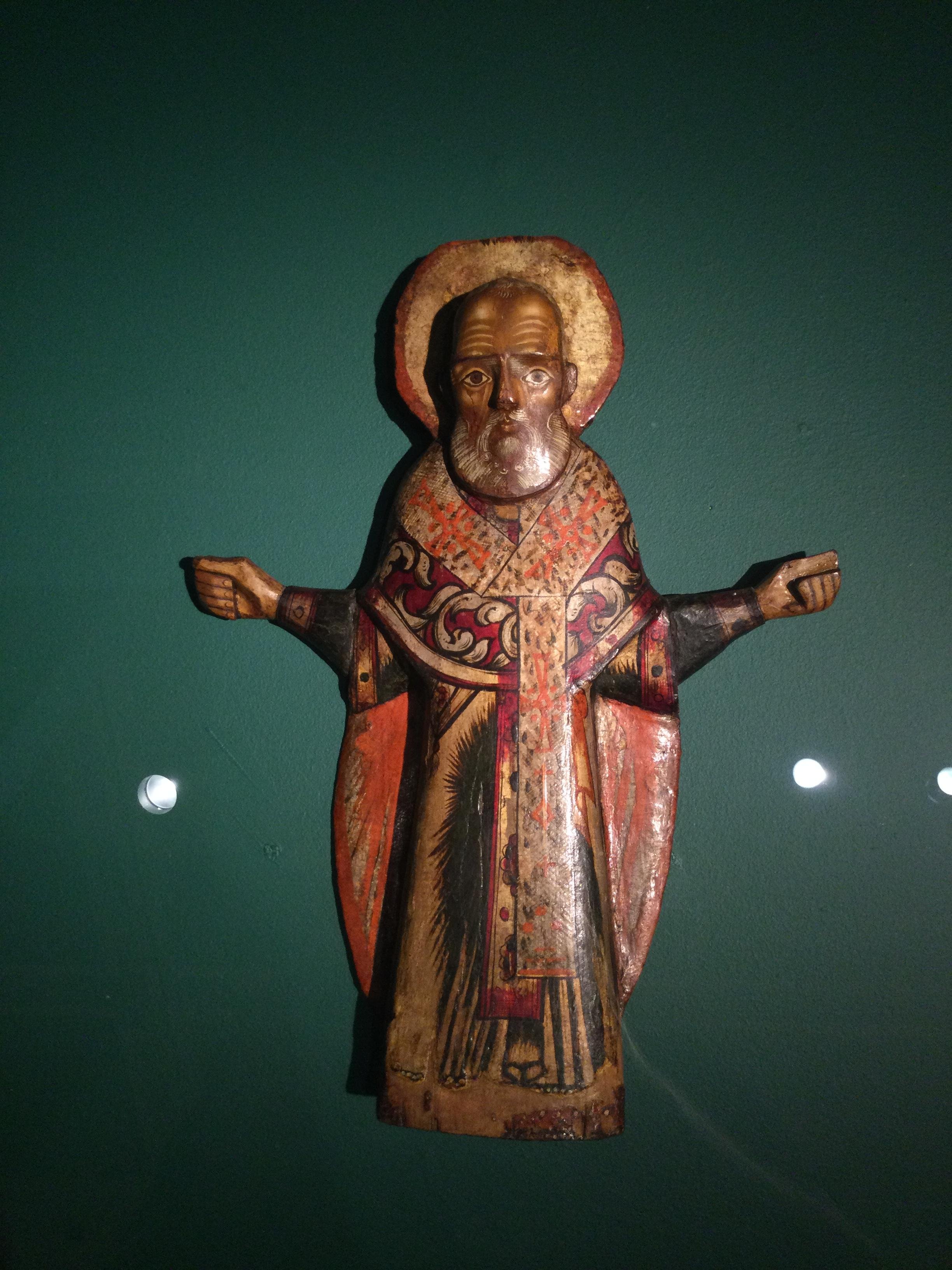 Religious Russian wooden sculpture of a st. Nicholas with arms outstretched, featuring a nimbus and detailed robes with red and gold patterns on a dark green background. Tretyakov gallery collection, Moscow