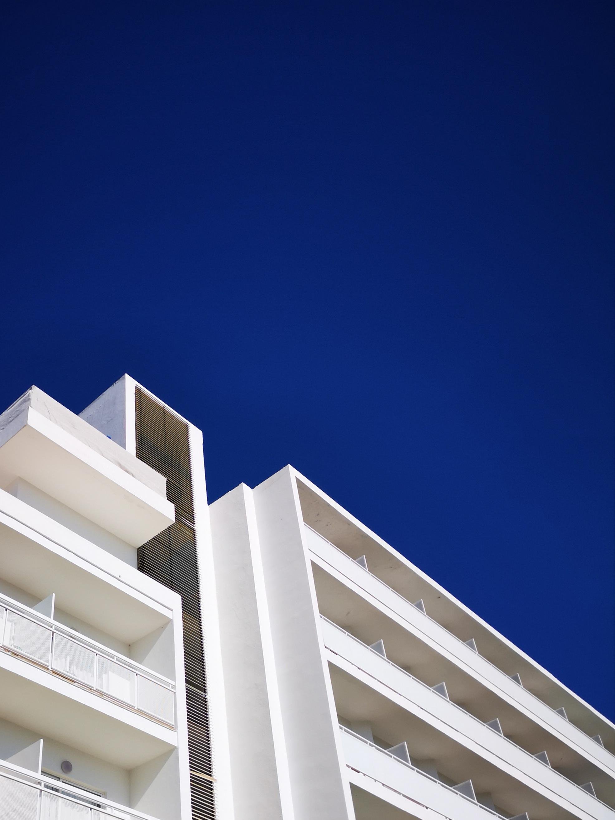 Upward view of a white modern apartment building with stacked balconies and railings, set against a deep blue sky.
