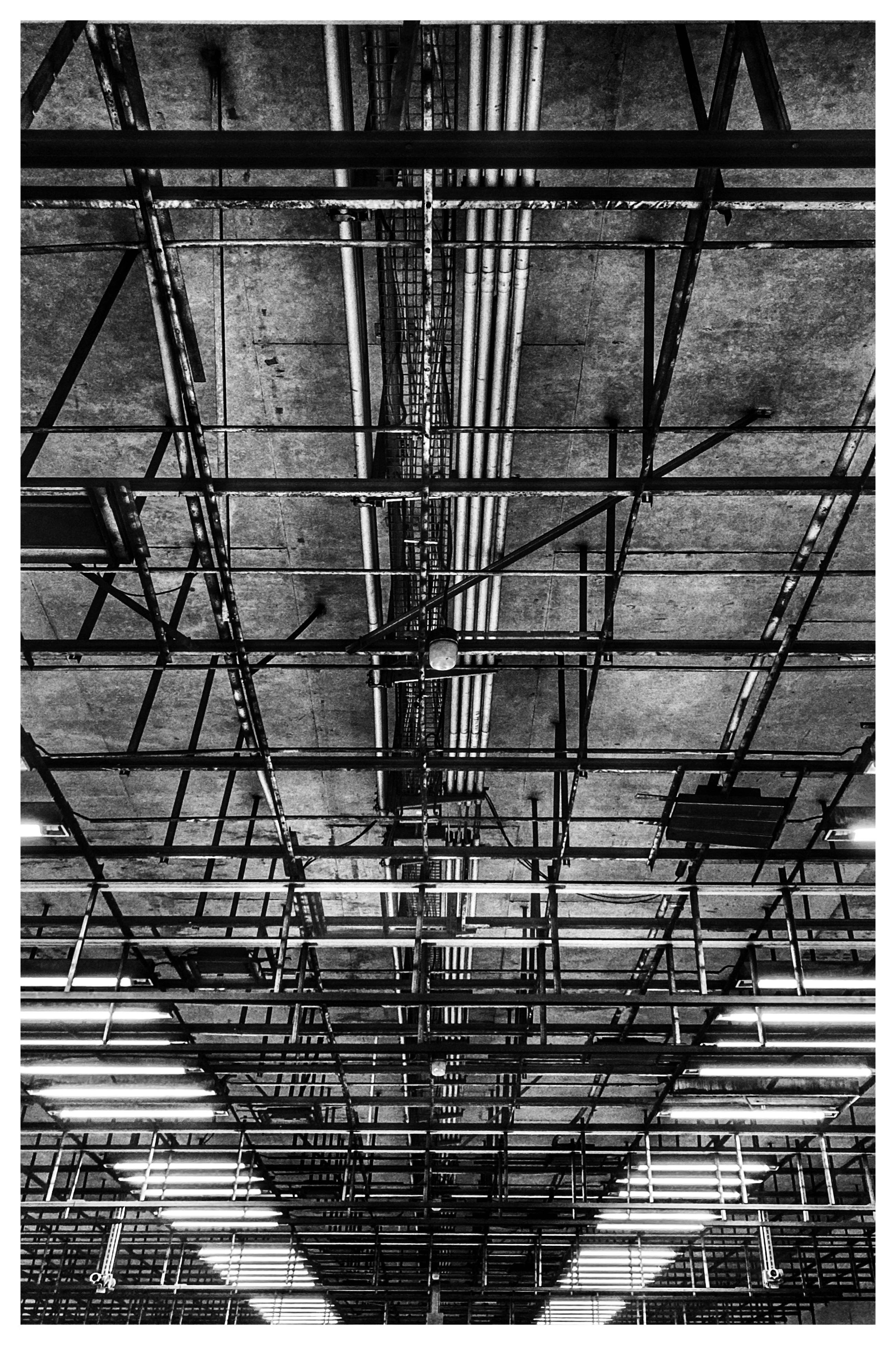 Upward view of an industrial ceiling with a grid of metal beams and scaffolding, centered bundles of pipes and cable trays, and rows of fluorescent lights near the bottom in a black-and-white photograph.
