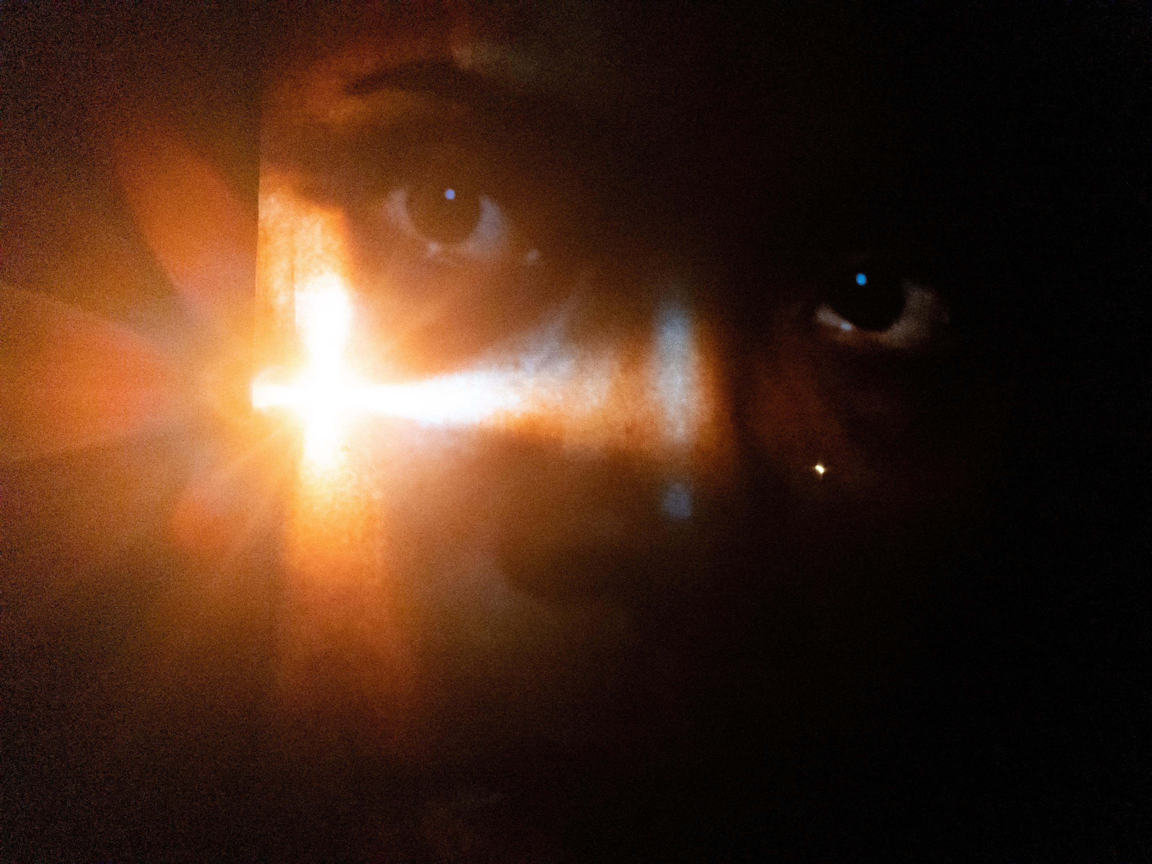 Close-up of a person's face partially illuminated by a bright, cross-shaped light. The light creates a lens flare effect and casts shadows, highlighting the eyes in the darkness.