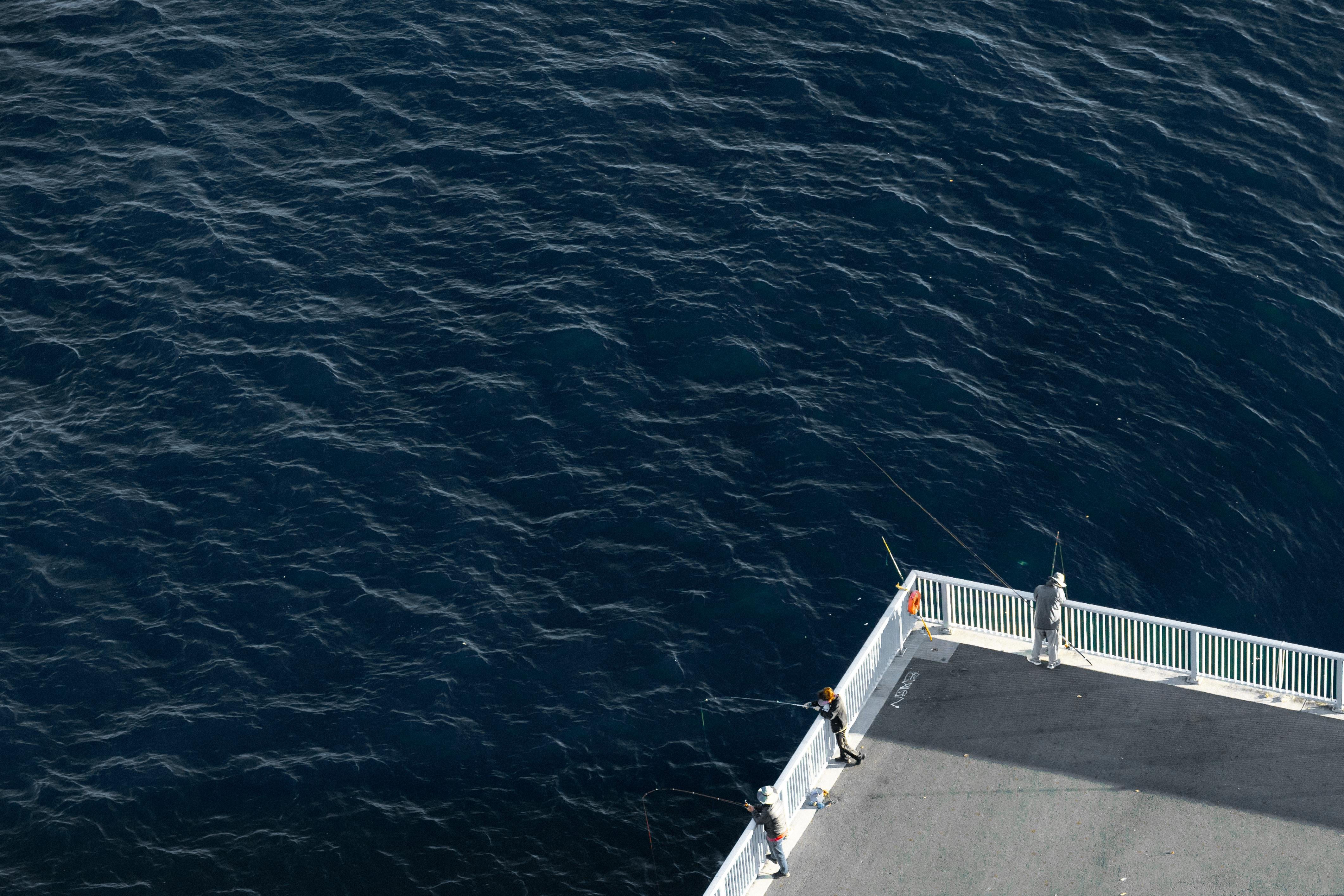 Aerial view of three people fishing from a fenced pier jutting out over a large expanse of deep blue water. The pier is angled diagonally across the lower right corner, with one person at the end and two others along the side. Shadows fall across the concrete surface, contrasting with the sunlight.