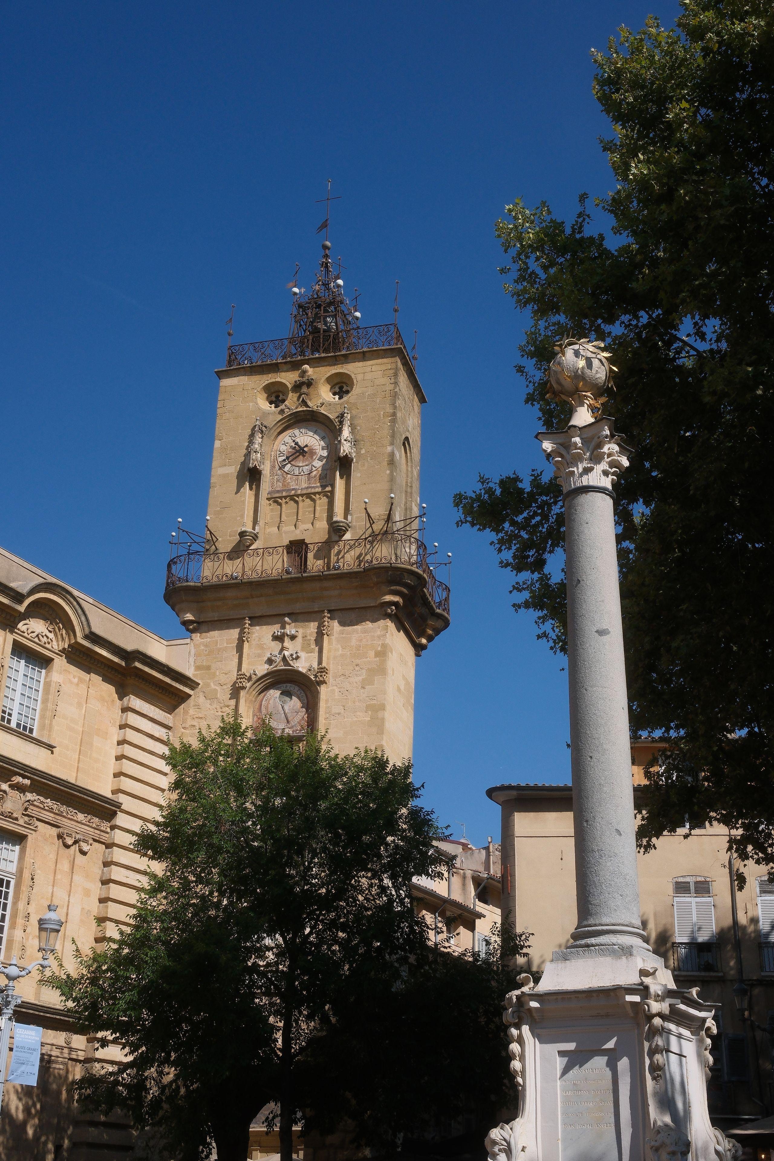 Photo format portrait avec au premier plan, à droite, un pilier ornemental avec un gros socle avec des moulures à chaque coins, et au sommet, une sphère ornée de feuilles d’or. À gauche, un clocher avec un chemin bordé d’une barrière, une horloge et au sommet avec la cloche. Sur fond de ciel bleu