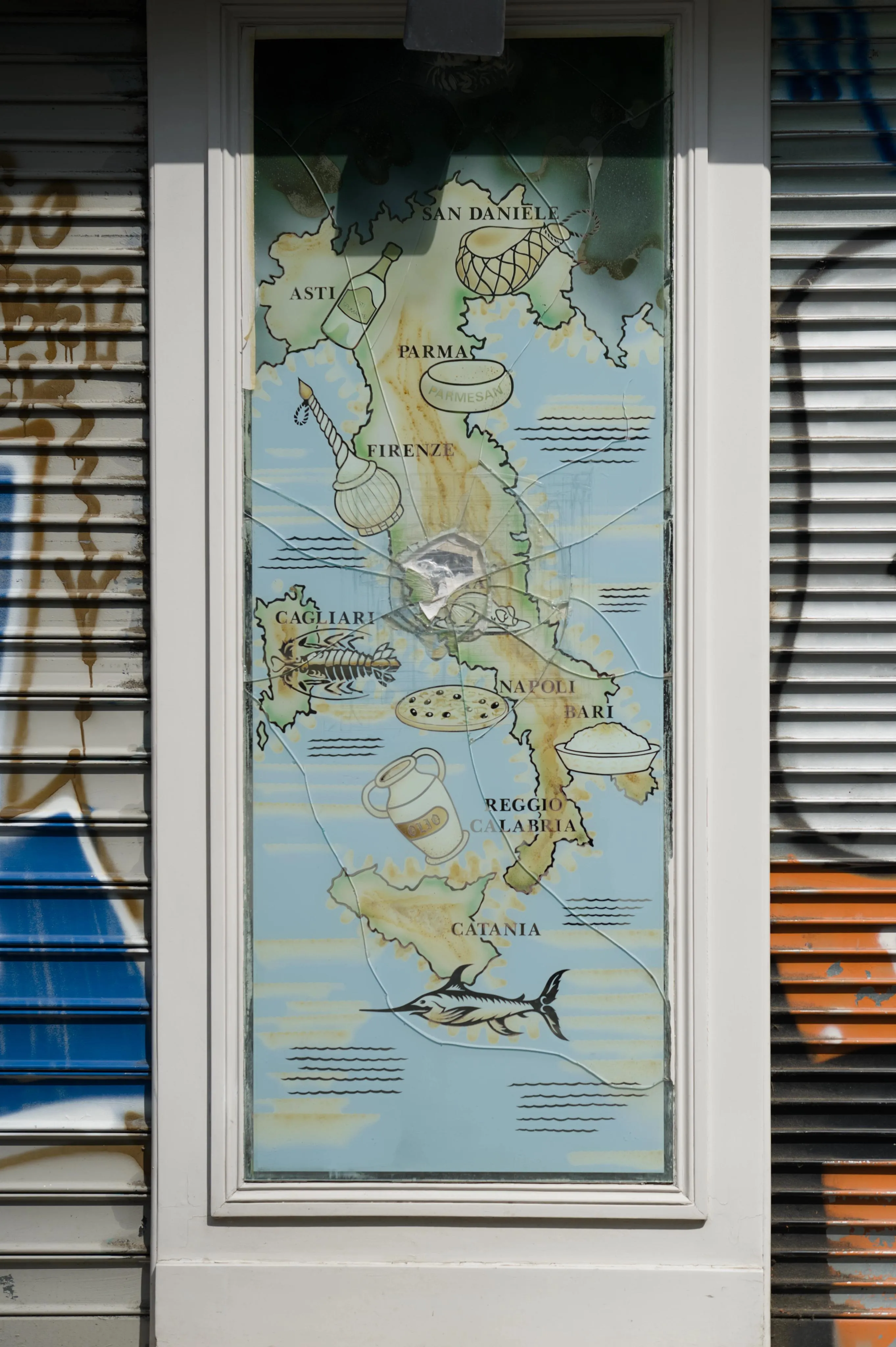Photograph of a cracked poster depicting the map of Italy on the wall of a shop in the street. Culinary specialties are represented around regions they are known for: pizza near Naples, parmesan near Parma among others.  The cracks in the protective glass over the poster are centered on the location of Rome, hiding the city and its label. Iron curtains surround the wall framing the poster on both sides.
