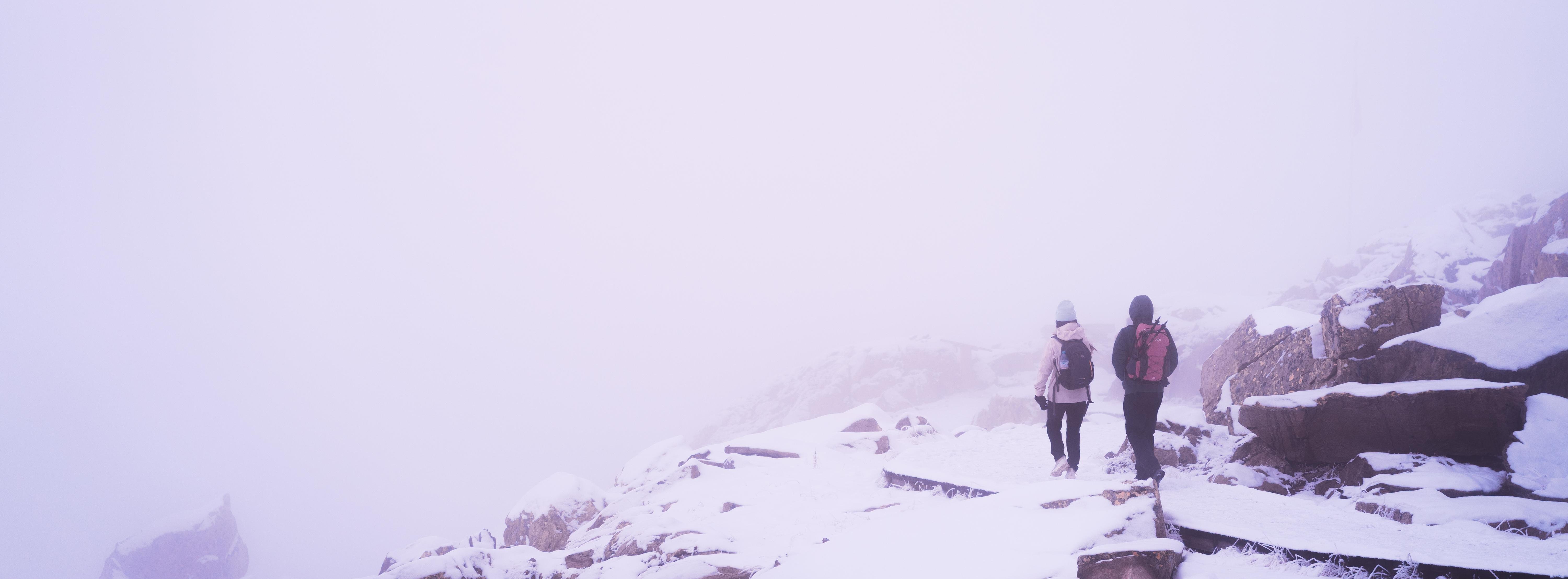 Two people dressed in winter clothing walk along a snow-covered, rocky path surrounded by dense fog. Jagged rocks border the pathway, and visibility is limited due to the misty atmosphere.