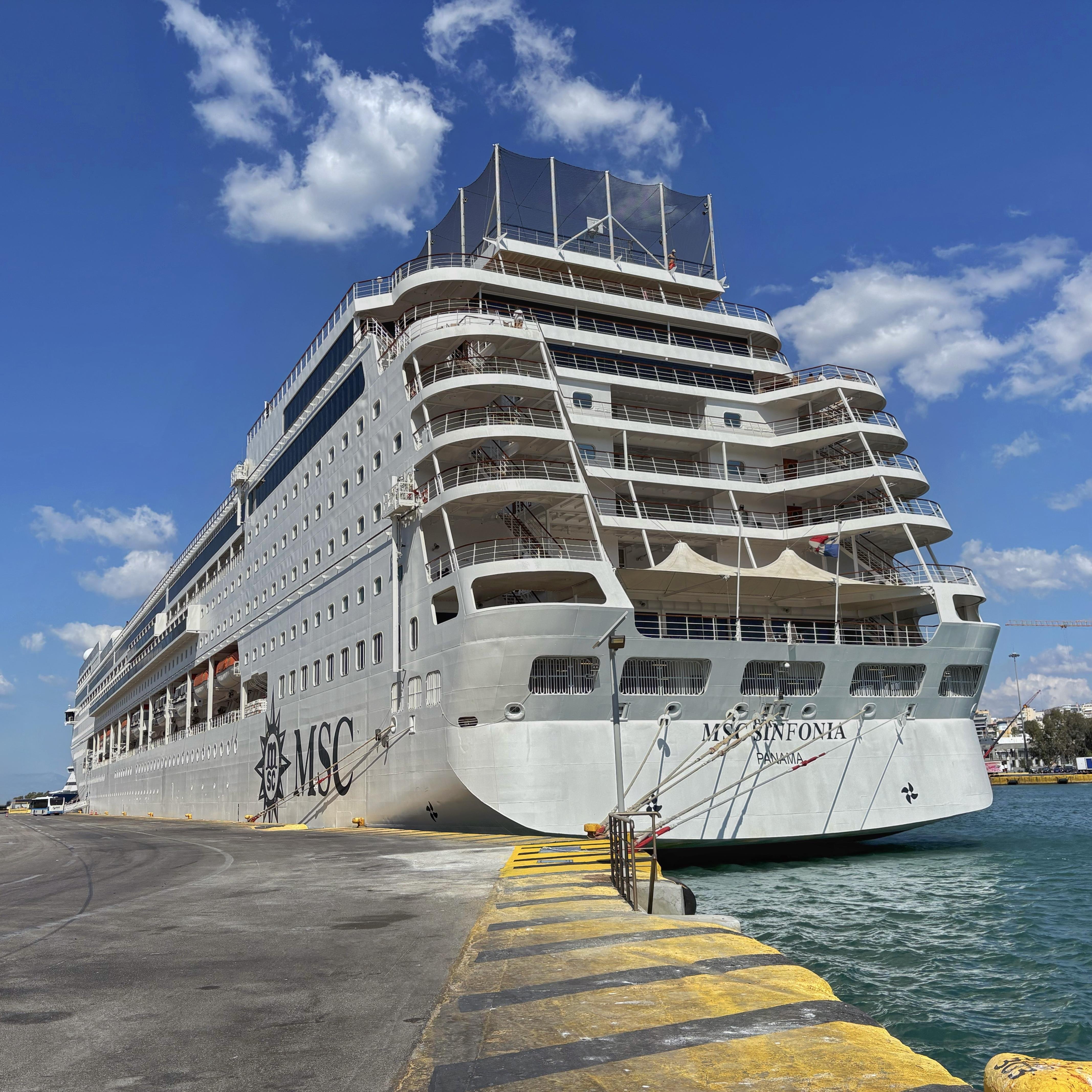 Large cruise ship docked at a port, featuring multiple decks and numerous windows. The ship displays the name "MSC Sinfonia" and flag details on the stern. The water is turquoise, and the sky is clear with some scattered clouds. A yellow-striped pathway is visible on the dock.