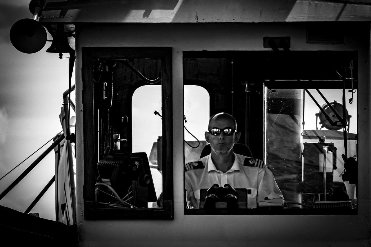 Monochrome image of a ship's captain, wearing a uniform with epaulettes, inside a control room. He is seated, facing forward, and wearing sunglasses. The room's interior features multiple windows, a steering apparatus, and various navigational equipment. Lighting and reflections add contrast, creating a defined and dramatic atmosphere.