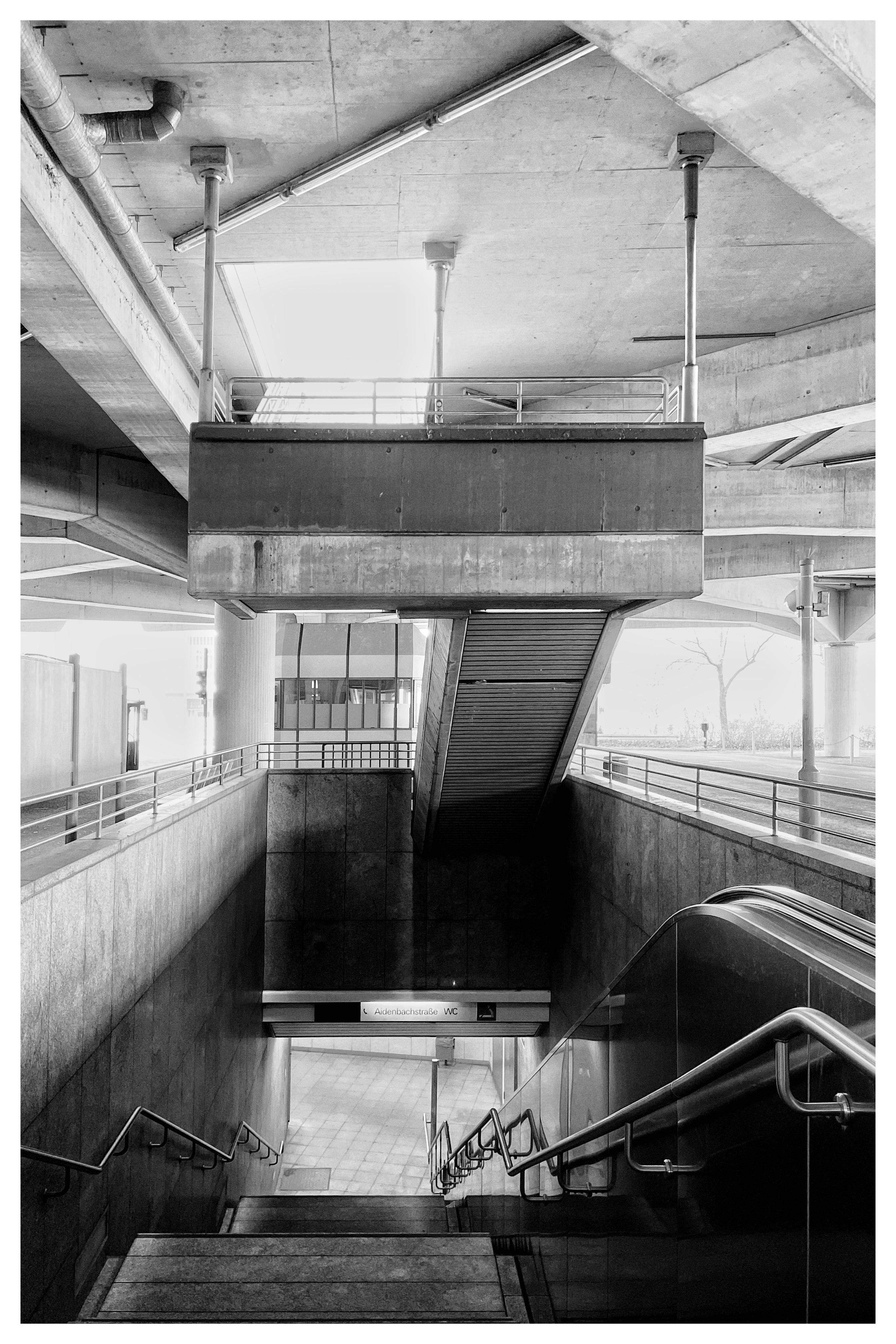 Black-and-white view down a concrete stairwell with metal handrails, leading to an underground transit entrance; an escalator runs along the right side, and overhead a concrete platform and beams with pipes and railings frame a bright opening.