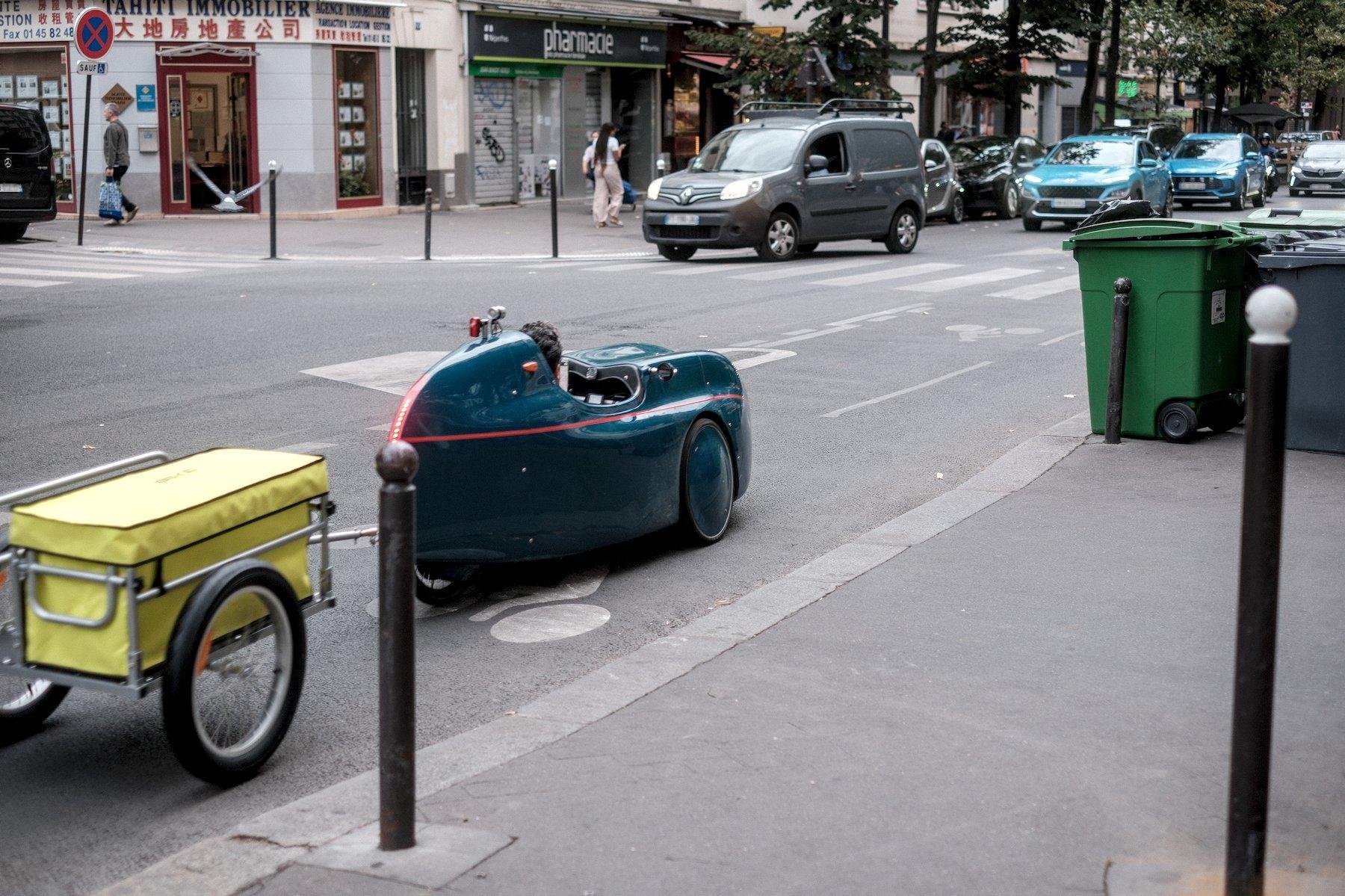 Photo horizontale. Sur une piste cyclable, un tricycle couché avec une carrosserie aérodynamique verte, tirant une petite remorque jaune