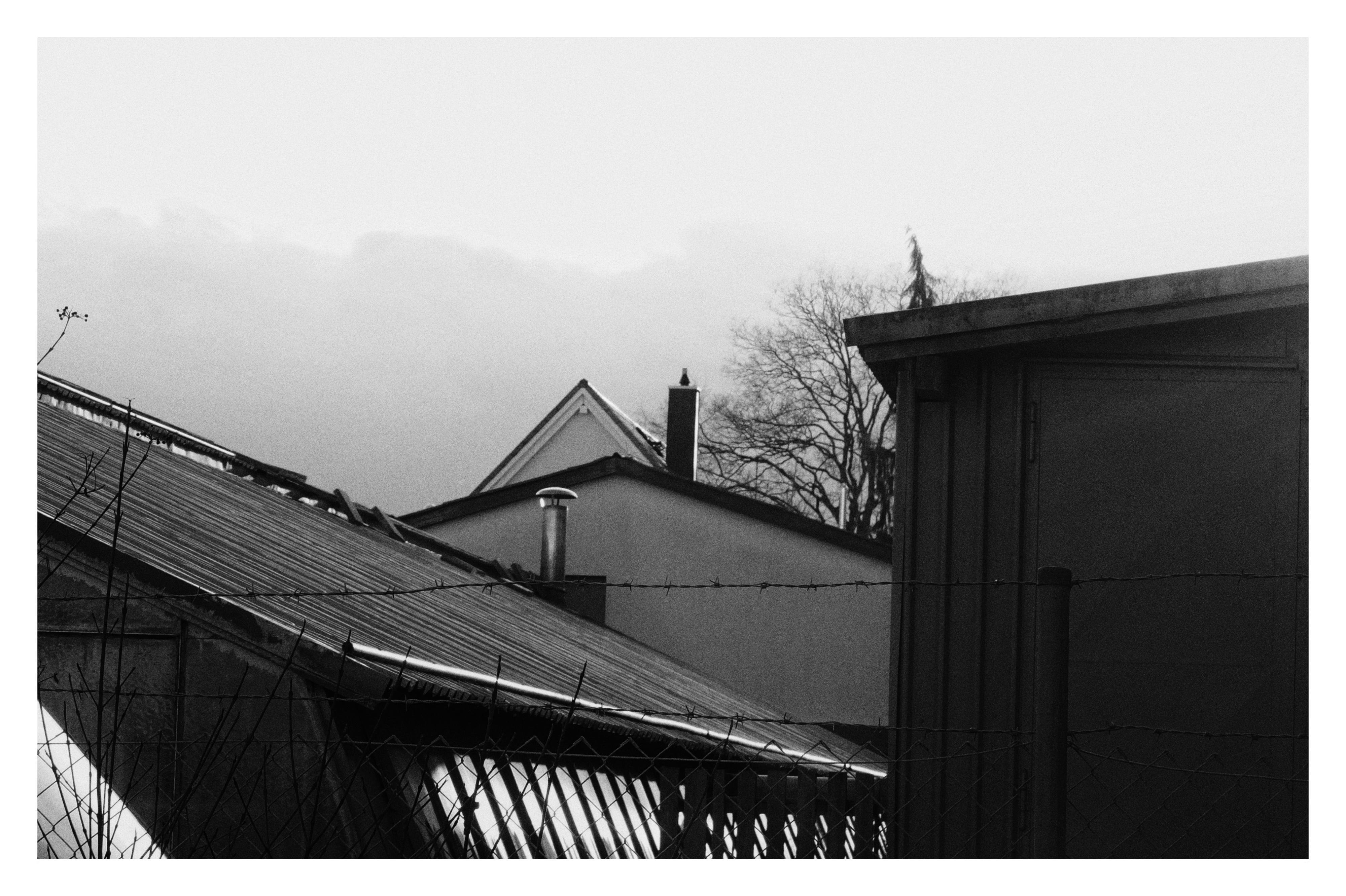 Black and white image of a residential area featuring rooftops of several buildings. A chimney and part of a tree are visible in the background. The foreground includes a fence with twisted wire, adding to the layered composition. Overcast clouds fill the sky above.