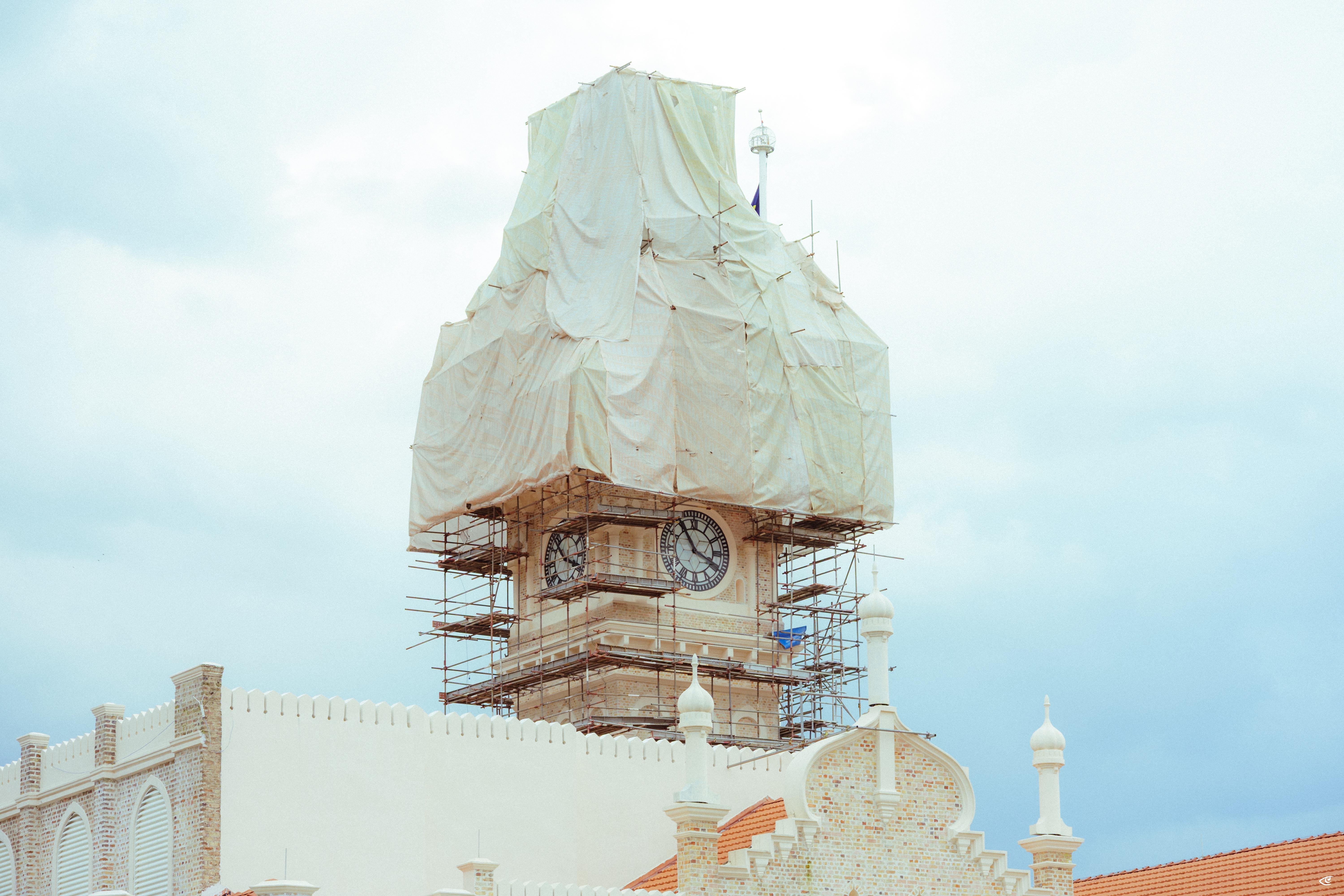 Clock tower under renovation with scaffolding and white tarps covering the upper section, rising behind an ornate white building with arched details against a cloudy sky.