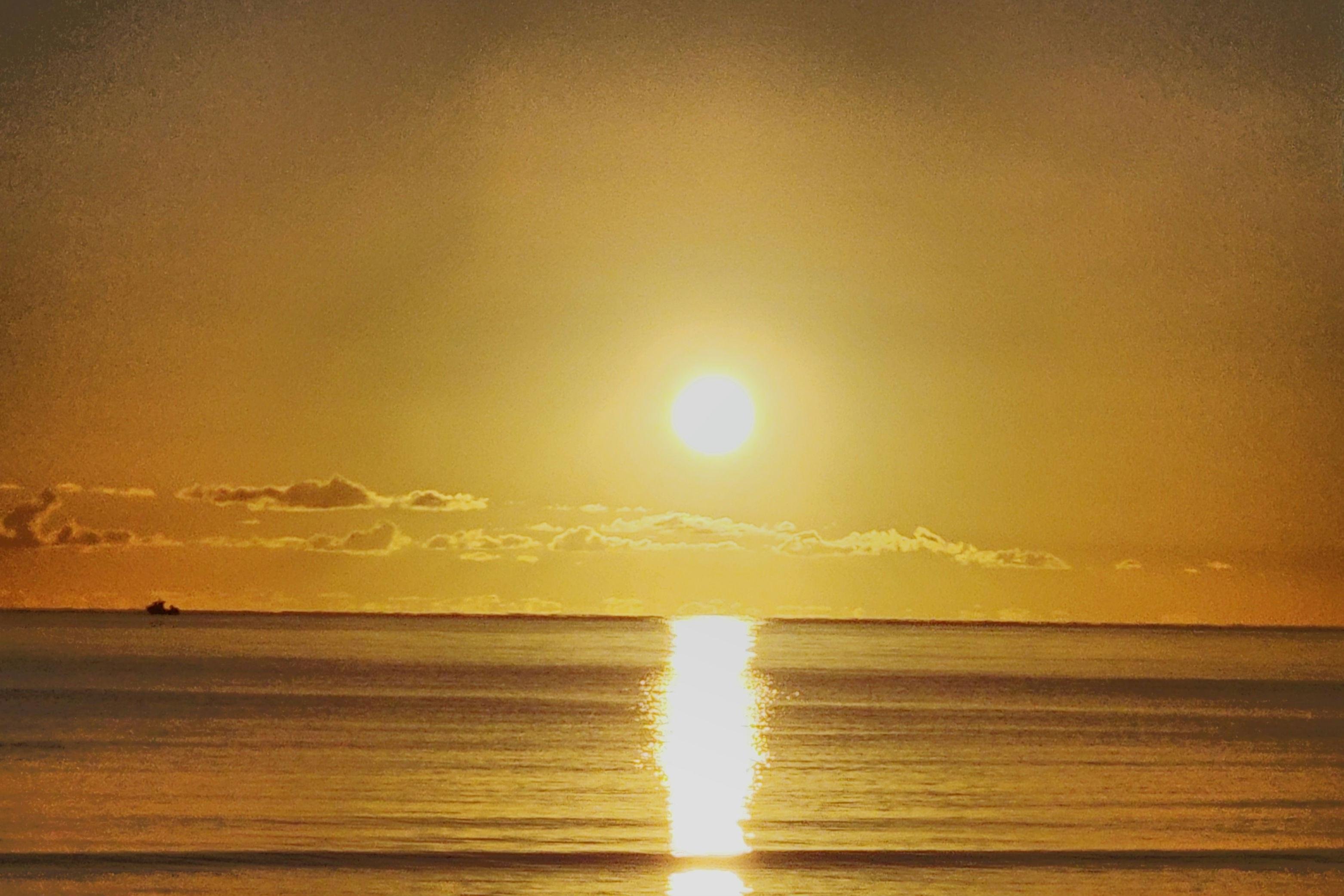 This image shows a sunrise over a calm body of water. The sun is low in the sky, creating a golden reflection on the water. There are some clouds on the horizon, and the overall tone of the image is warm and serene.