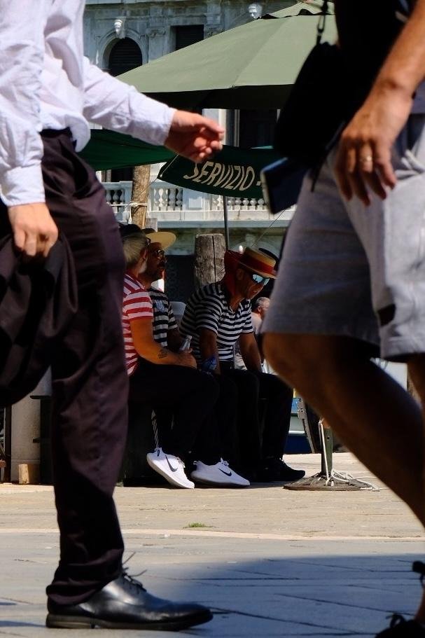 Three Venetian Gondola drivers sitting in the shade of a green umbrella waiting for customers. On the umbrella yellow letters spell the word „SERVIZIO“.

You can see the scene through the legs of pedestrians passing by.