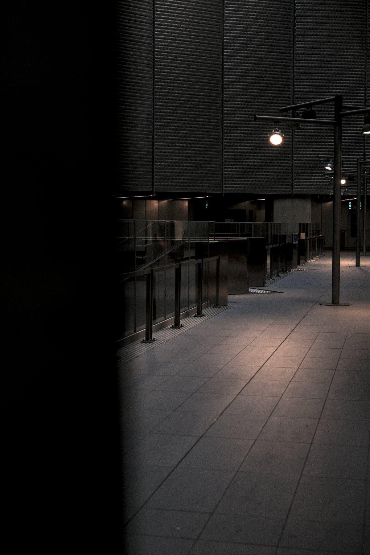Photo verticale. Vue derrière un mur ou un poteau, à gauche, sur un passage désert avec des lampadaires projettant des tâches de lumières blafardes sur le sol carrelé