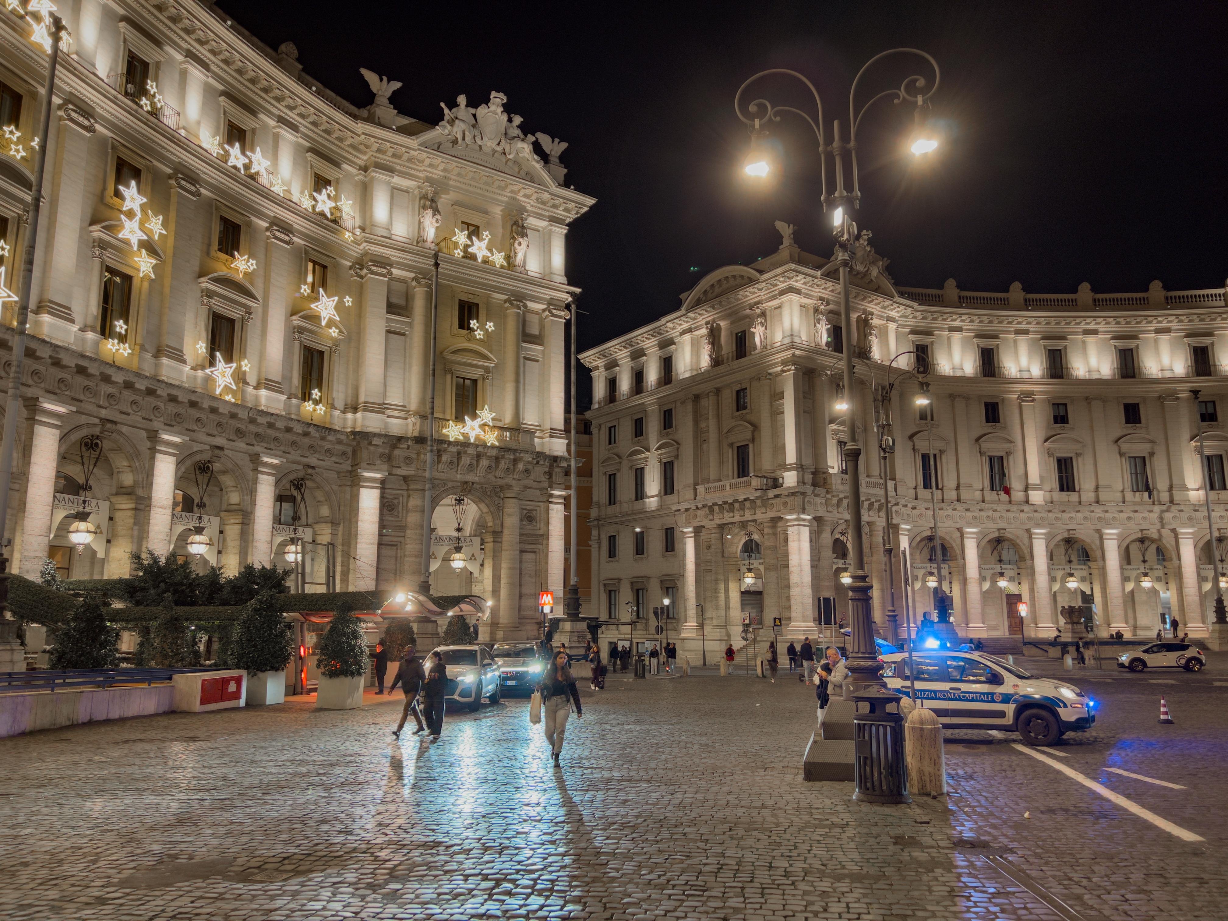 Scena notturna di una piazza urbana splendidamente illuminata con architettura classica ornata. Gli edifici sono adornati con luci decorative e ci sono pedoni che camminano lungo la piazza pavimentata con ciottoli. C'è una macchina della polizia con le luci accese, parcheggiata sul lato destro, e un lampione che illumina l'area. L'ambientazione suggerisce una città europea, forse durante una stagione festiva.