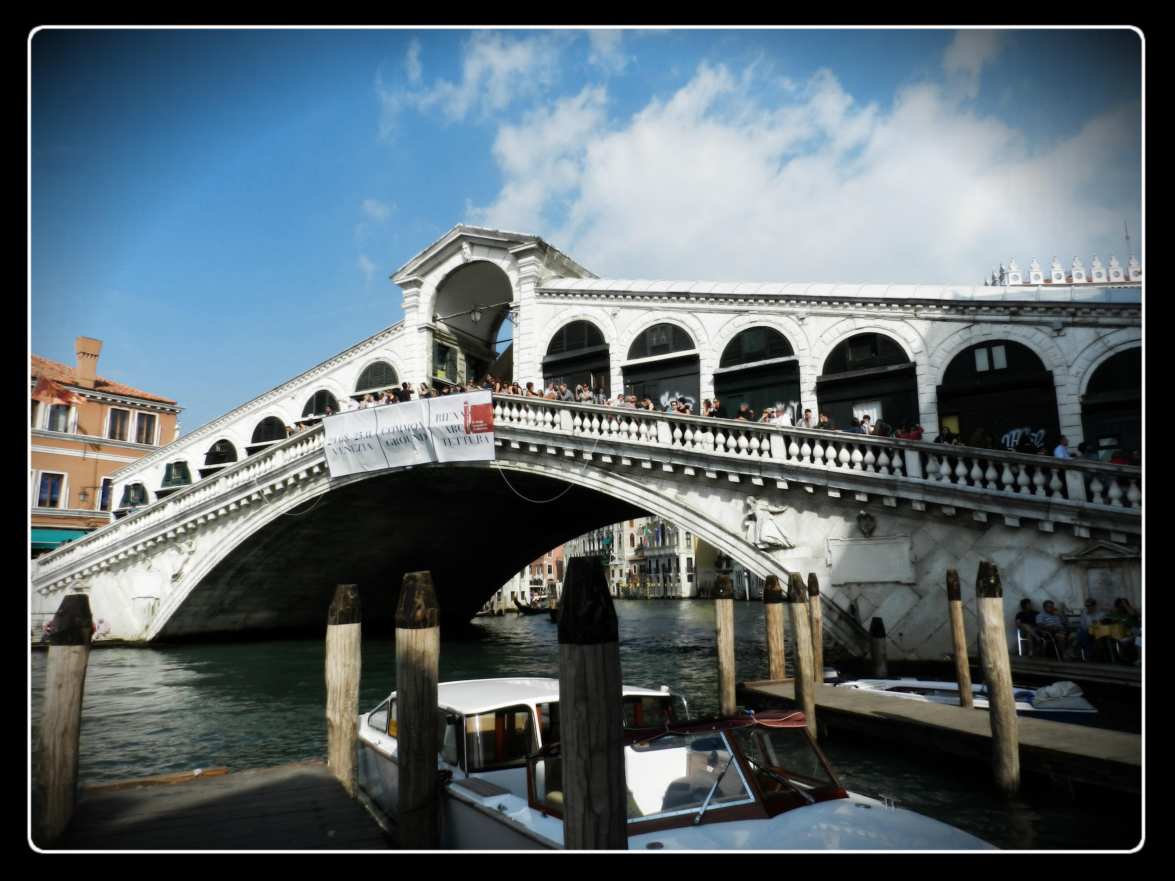 Photograph of the iconic Ponte di Rialto spanning the Grand Canal in Venice, Italy, taken from a low angle near the water’s edge. The elegant white-stone bridge, completed in 1591 during the Renaissance, features a single sweeping arch with rows of rounded arches lining both sides. Under the bright blue sky, crowds of tourists can be seen walking along the central walkway and browsing the small shops built directly into the bridge—one of its most distinctive features. A large banner hangs along the balustrade, partially shading the stone. Below, water taxis and private boats are moored at wooden posts typical of Venetian canals. The canal stretches into the distance beneath the arch, framed by colorful historic buildings on either side. The scene captures the lively, bustling atmosphere around Venice’s oldest and most famous bridge, celebrated for its architectural brilliance and its longstanding role as a central crossing point of the Grand Canal.