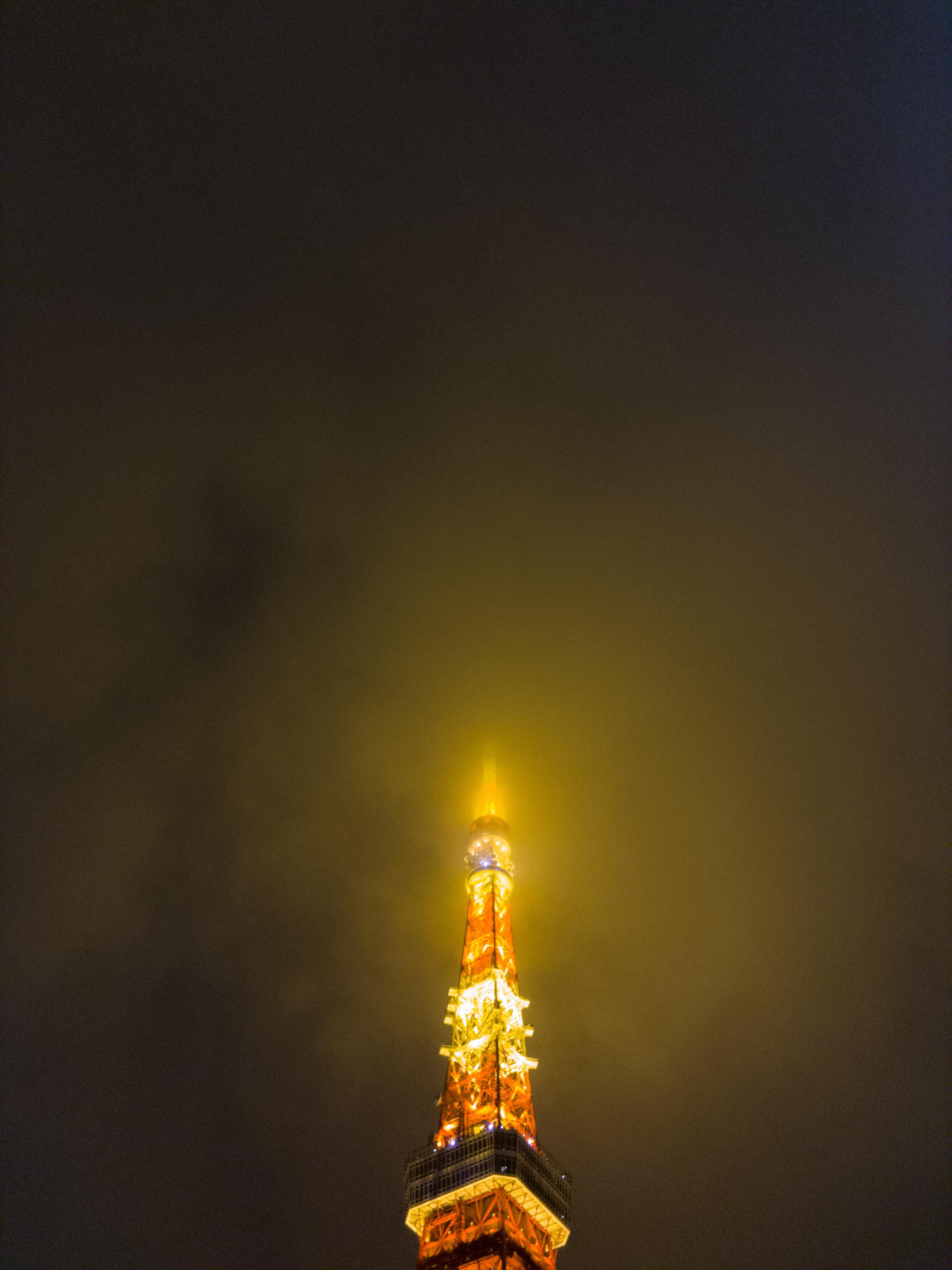 Orange and white illuminated tower structure piercing through a dark, cloudy night sky with a glowing effect at the top.