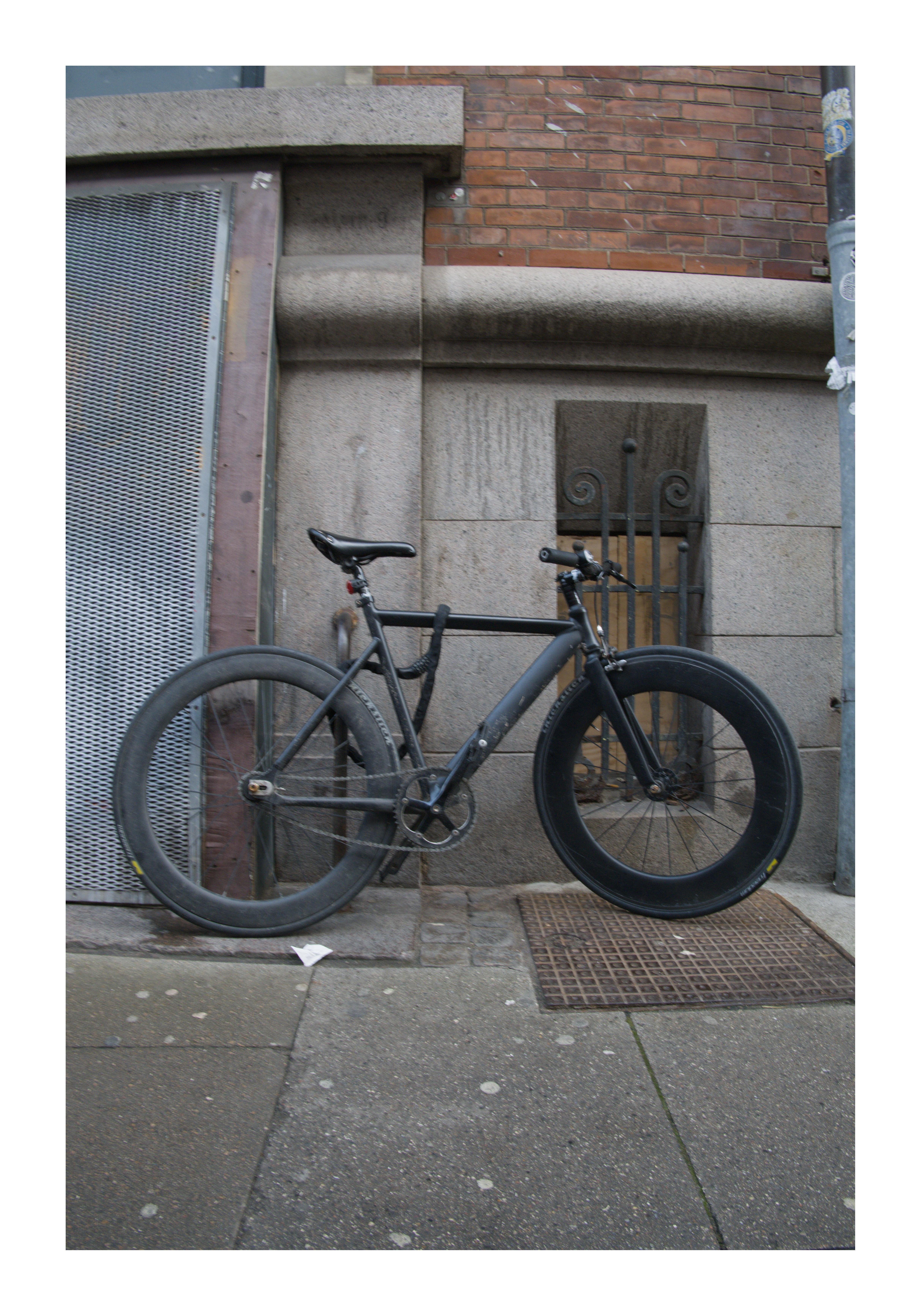 Black bicycle with a deep-section front wheel leaning against a stone-and-brick building wall; metal vented door on the left, barred window behind the bike, and a sidewalk with a grated utility cover in the foreground.