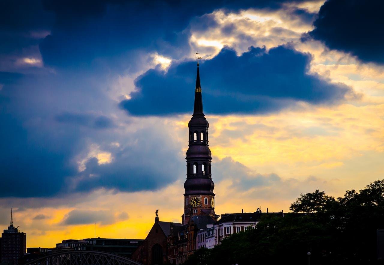 Tall church steeple with a clock tower rising above low buildings, silhouetted against a yellow-orange sunset sky with large dark clouds.