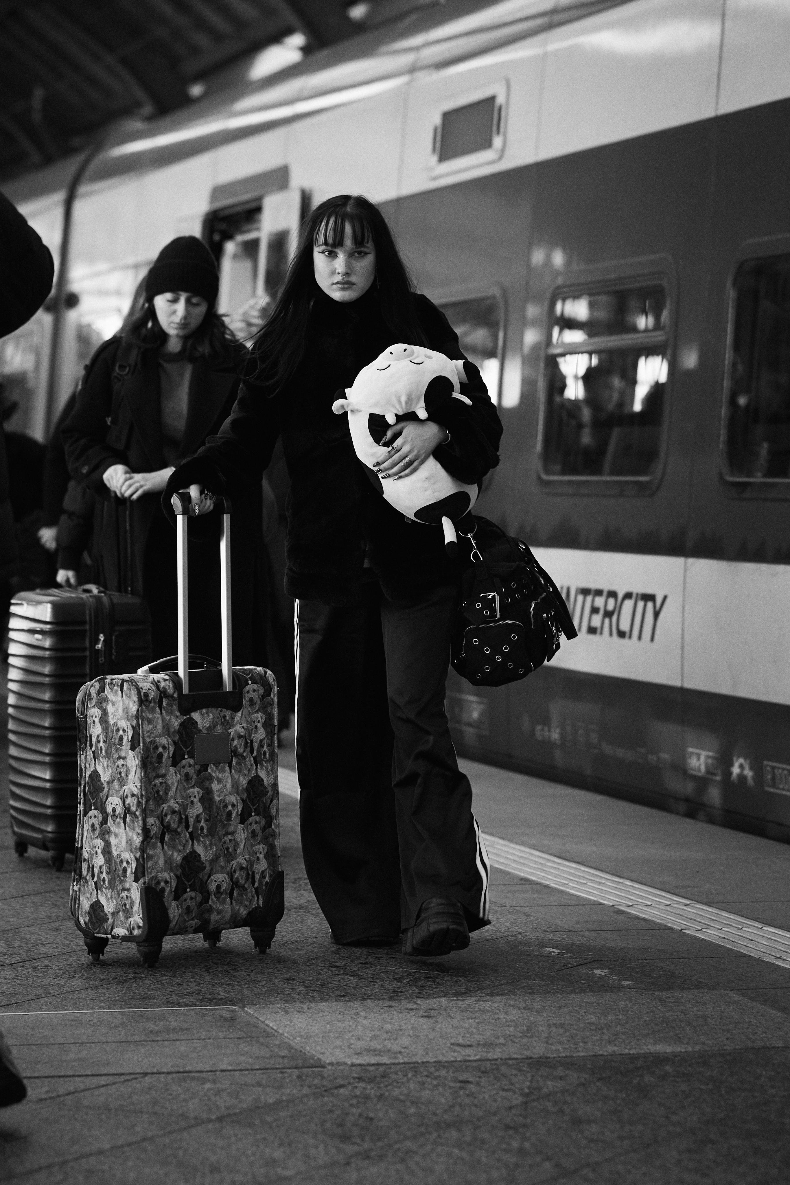 The image shows a black and white scene at a train station. A person is holding a plush toy and pulling a suitcase with a dog pattern. Another person in the background is pulling a suitcase as well. The train has the word "INTERCITY" visible on it.