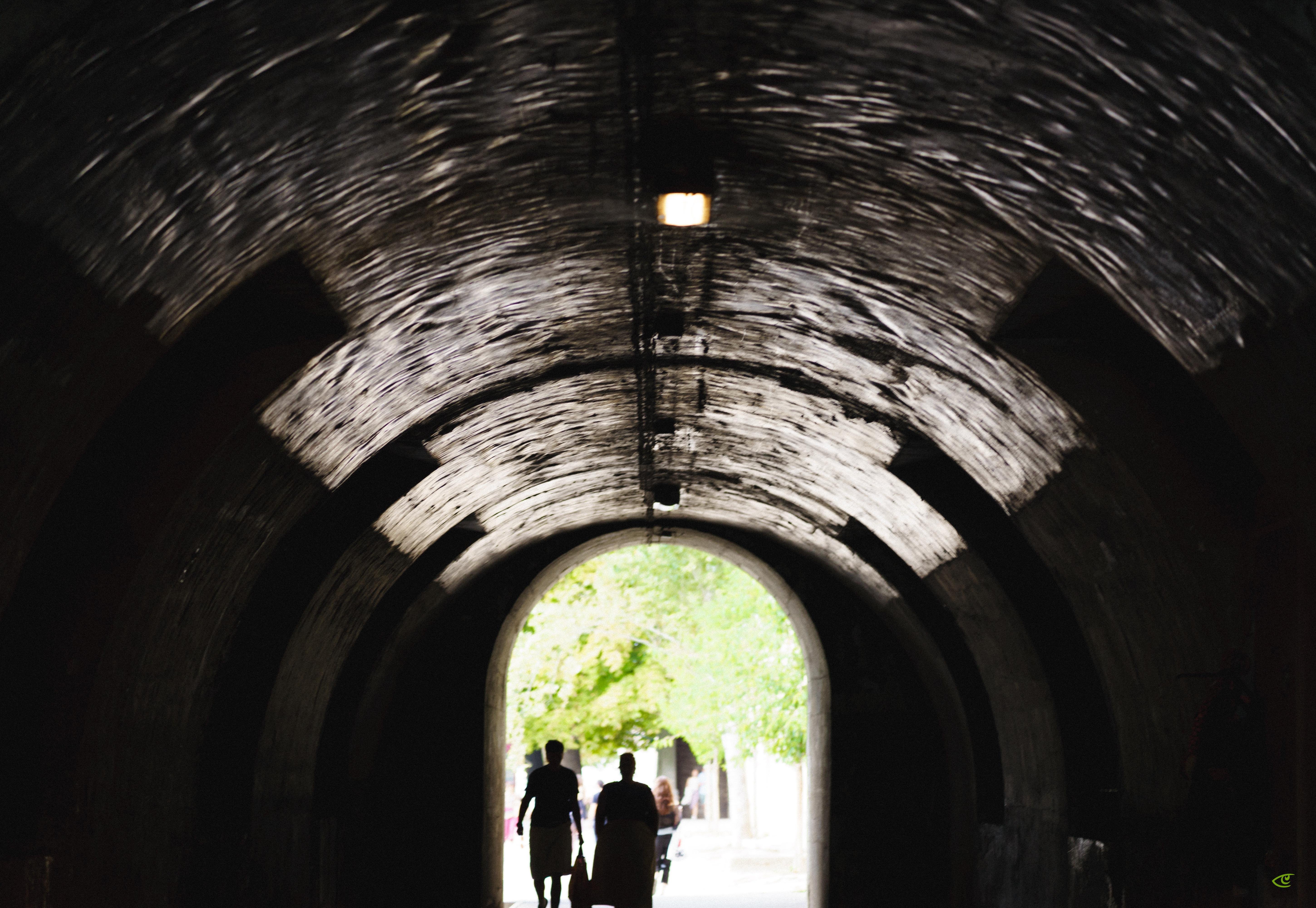 Silhouettes of two people walking through a dark, arched tunnel toward a bright exit with green trees visible outside; curved tunnel ceiling with a single light fixture overhead.
