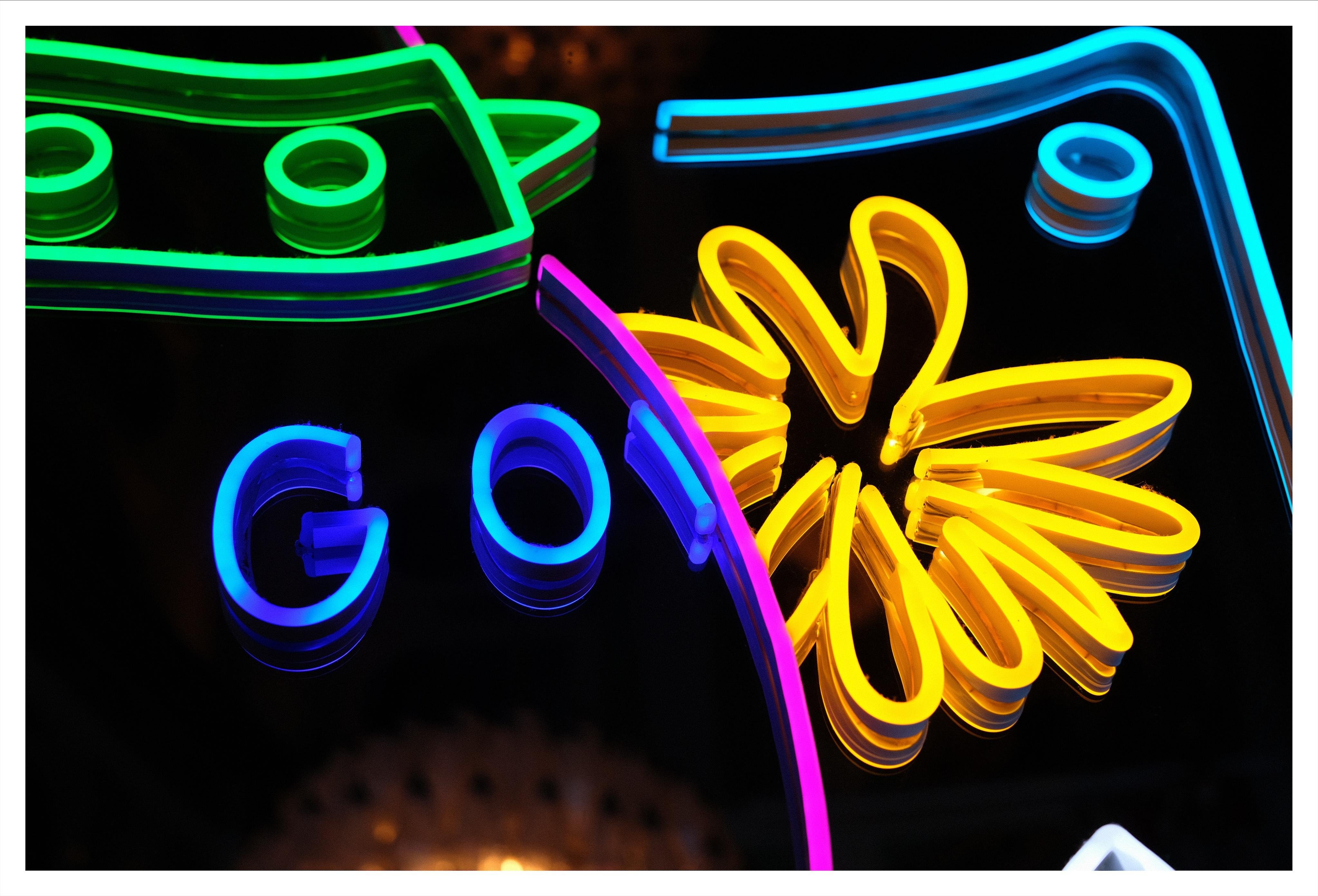 Close-up of a neon sign on a dark mirror, with blue letters “GO,” a yellow flower-like burst shape, and curved green, purple, and blue neon shapes.