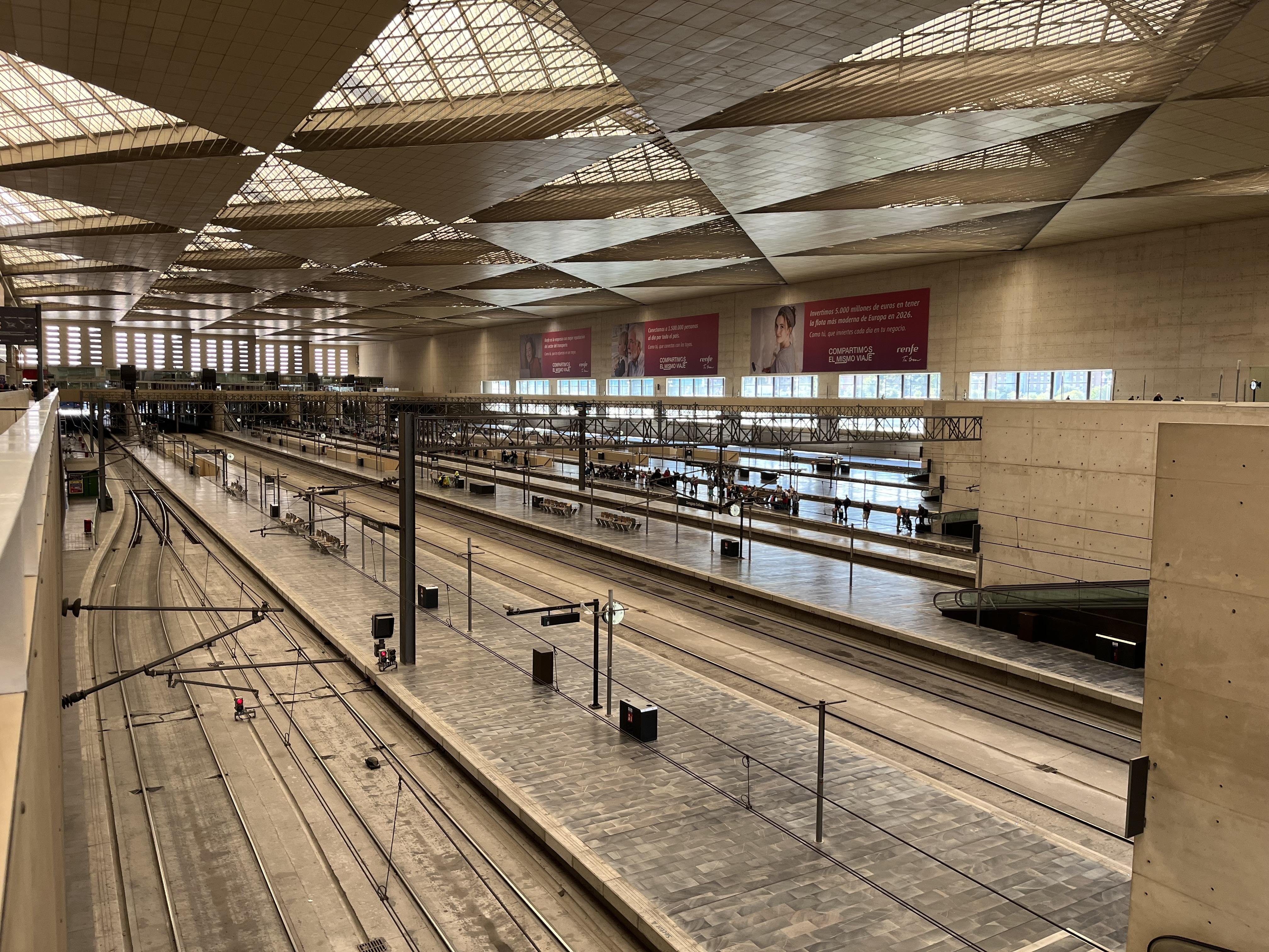 The image shows a large, modern train station with multiple railway tracks and platforms. The interior features a spacious design with a high ceiling and ample natural light from large windows. There are overhead structures for power lines and signage. The station appears empty, with no trains or passengers visible.