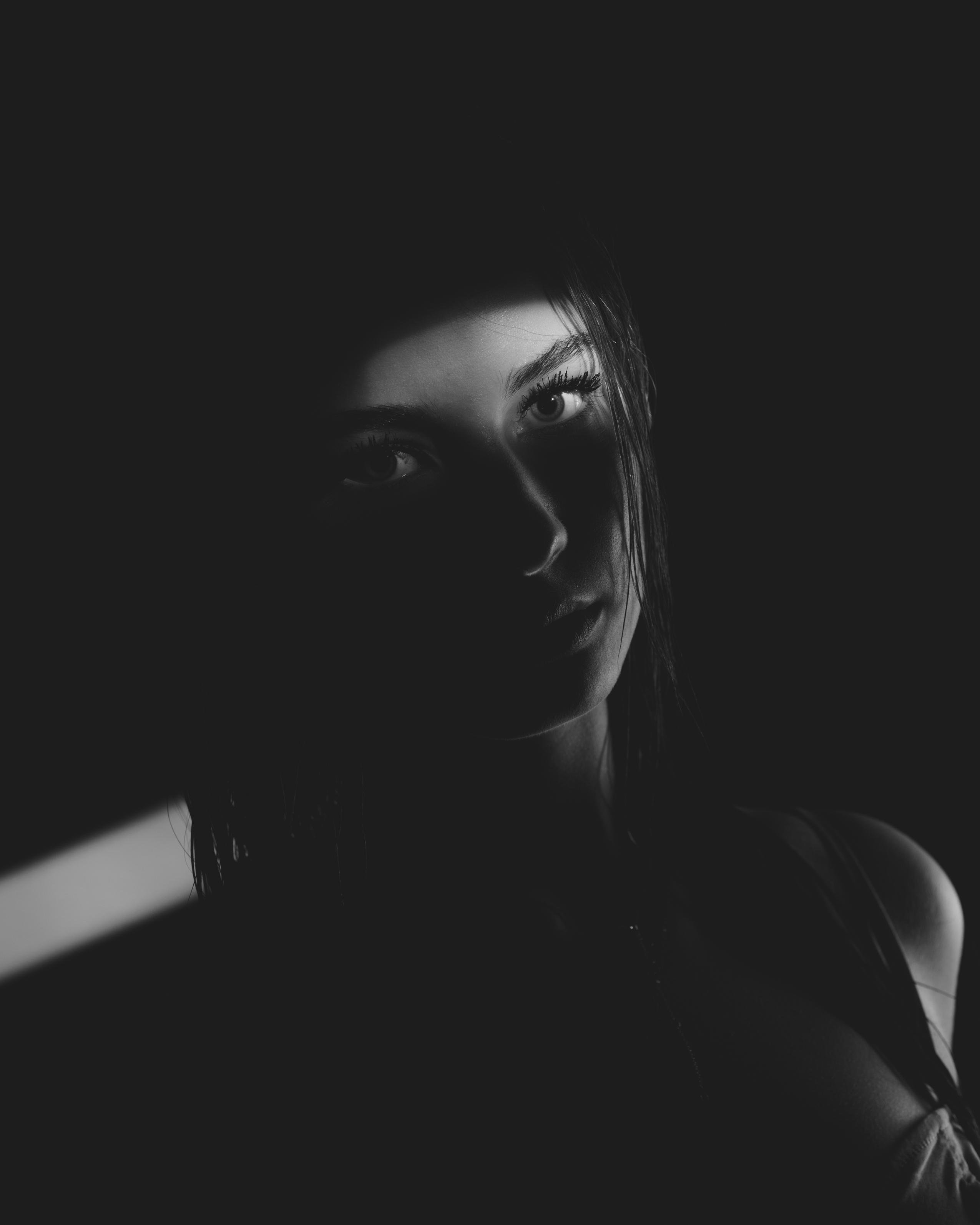 A black and white portrait of a woman in low light with one side of her face partially illuminated, creating strong contrast. Her eyes and partial silhouette are visible, while the rest of her face recedes into shadows. Light enters from the left side, adding a dramatic effect.