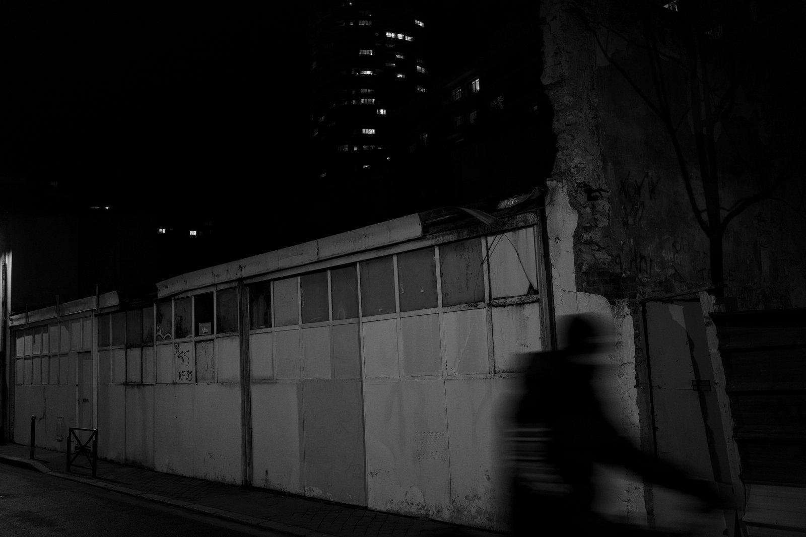Photo horizontale, noir et blanc, de nut. Une façade blanche sale d’un petit bâtiment sans étage, baricadé et qui semble abandonné. Sur la droite, la silhouette floue d’une personne passant à vélo. En haut, un fond noir avec des petite lumière émanant de fenêtre d’un grand immeuble