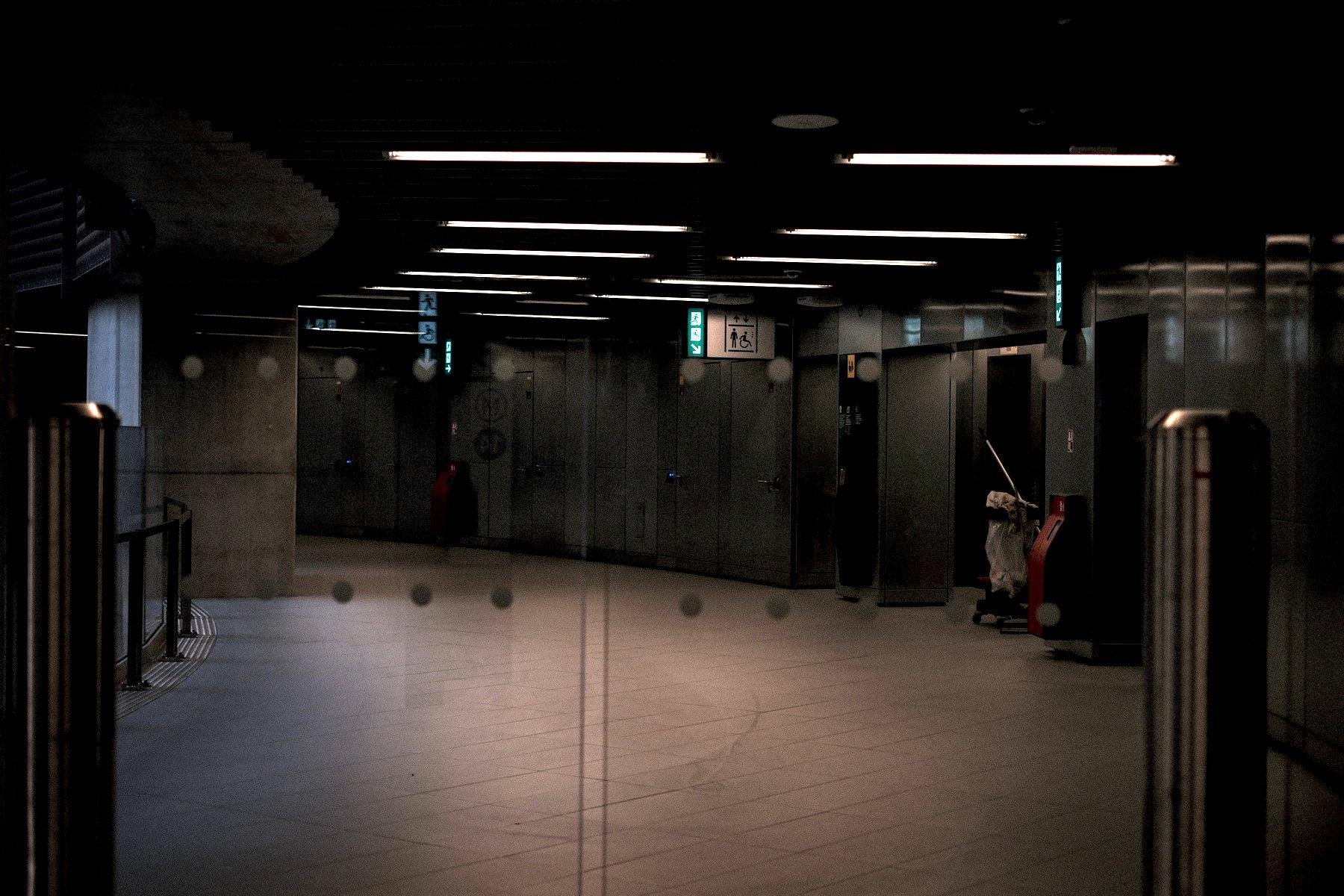 Photo horizontale. Couloir courbé de métro, derrière un portail en verre, les néons parallèles suivant la courbure du couloir, éclairent le vide dérangé par un chariot d’entretiens de propreté