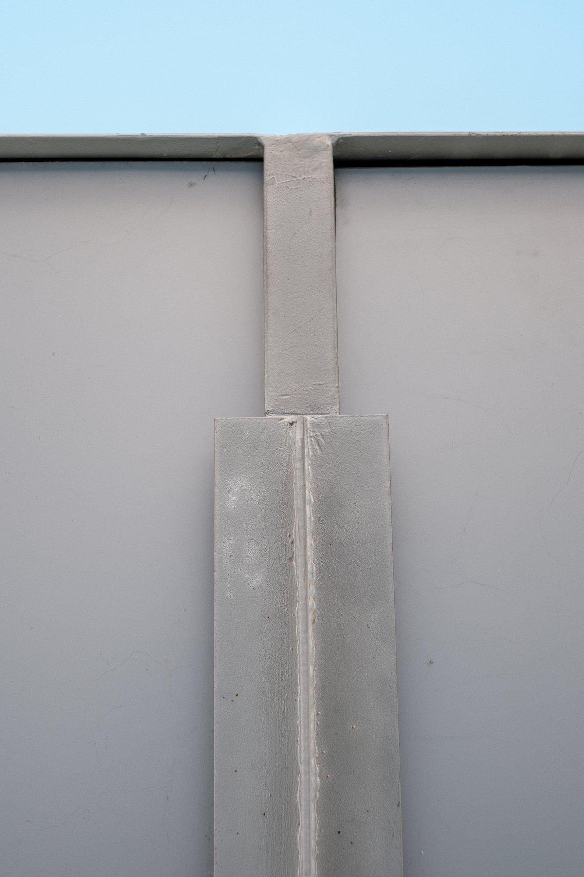 Photo verticale. Tout en haut, un bout de ciel tout bleu clair, sur environ ⅙ de l’image, le reste est occupé par le dessous d’un toit gris, avec une barre de soutiens. L’ensemble est très abstrait