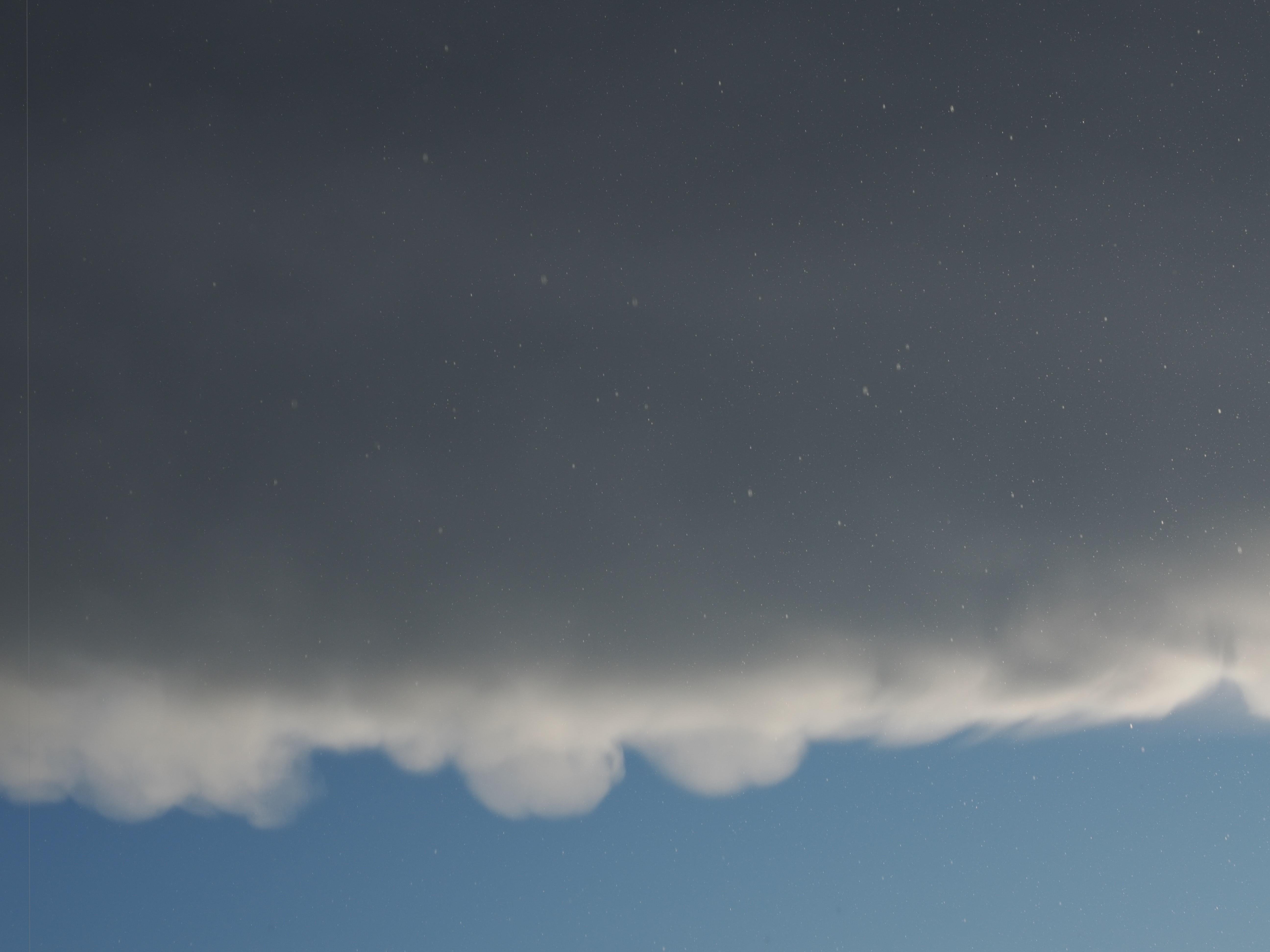 Dark gray clouds cover the upper two-thirds of the image with a distinct, wavy edge separating them from the clear blue sky below. Scattered, small white specks like raindrops are visible throughout the sky.