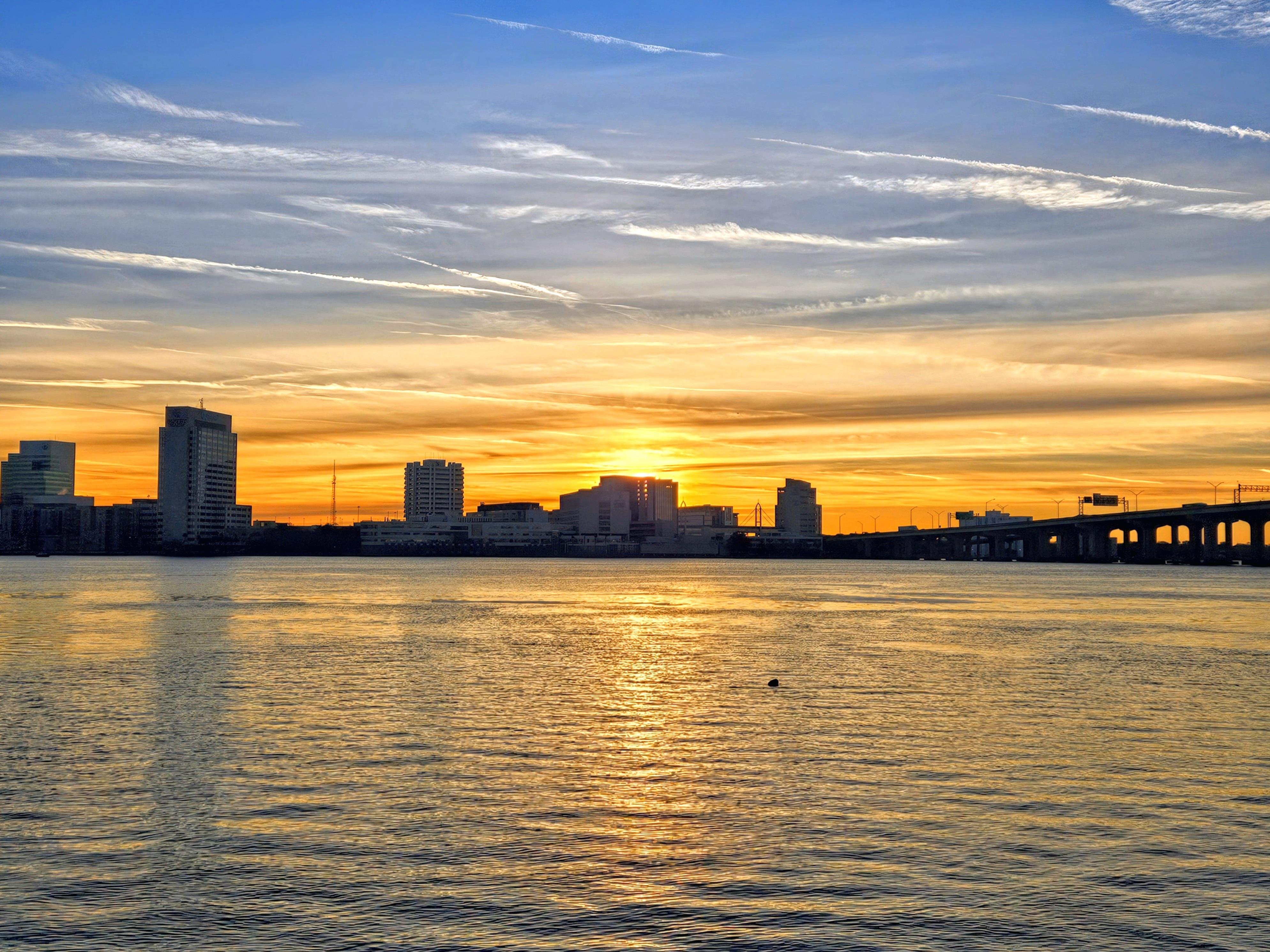 Sunrise over a city skyline with several tall buildings silhouetted against a vibrant sky. The sun is partially hidden behind the buildings, casting a golden reflection on the water in the foreground. A bridge is visible on the right side, stretching across the scene. Wispy clouds streak the sky, transitioning from blue to warm orange hues.