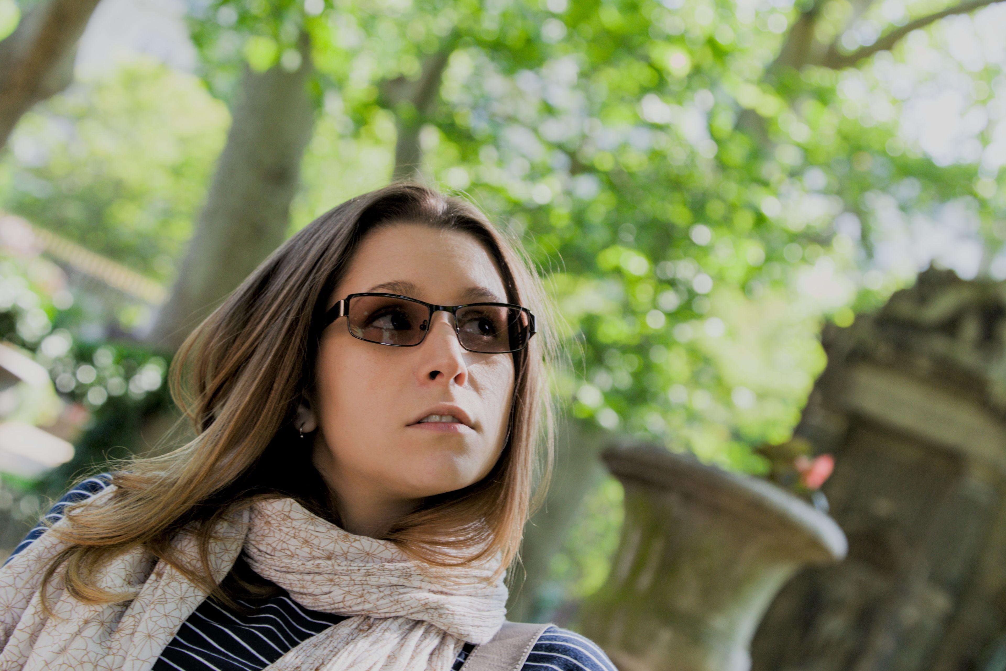 Photo horizontale. une jeune femme brune au cheveux long, portant un haut bleu marine à rayure horizontales blanches, un foulard blanc au motifs floraux marron, des lunettes auto teintées, penchant la tête d’un côté et regardant en l’air de l’autre côté. Le fond est verdoyant avec un pot de fleur en pierre teinté de mousse. Le cadrage est aussi penché vers la droite